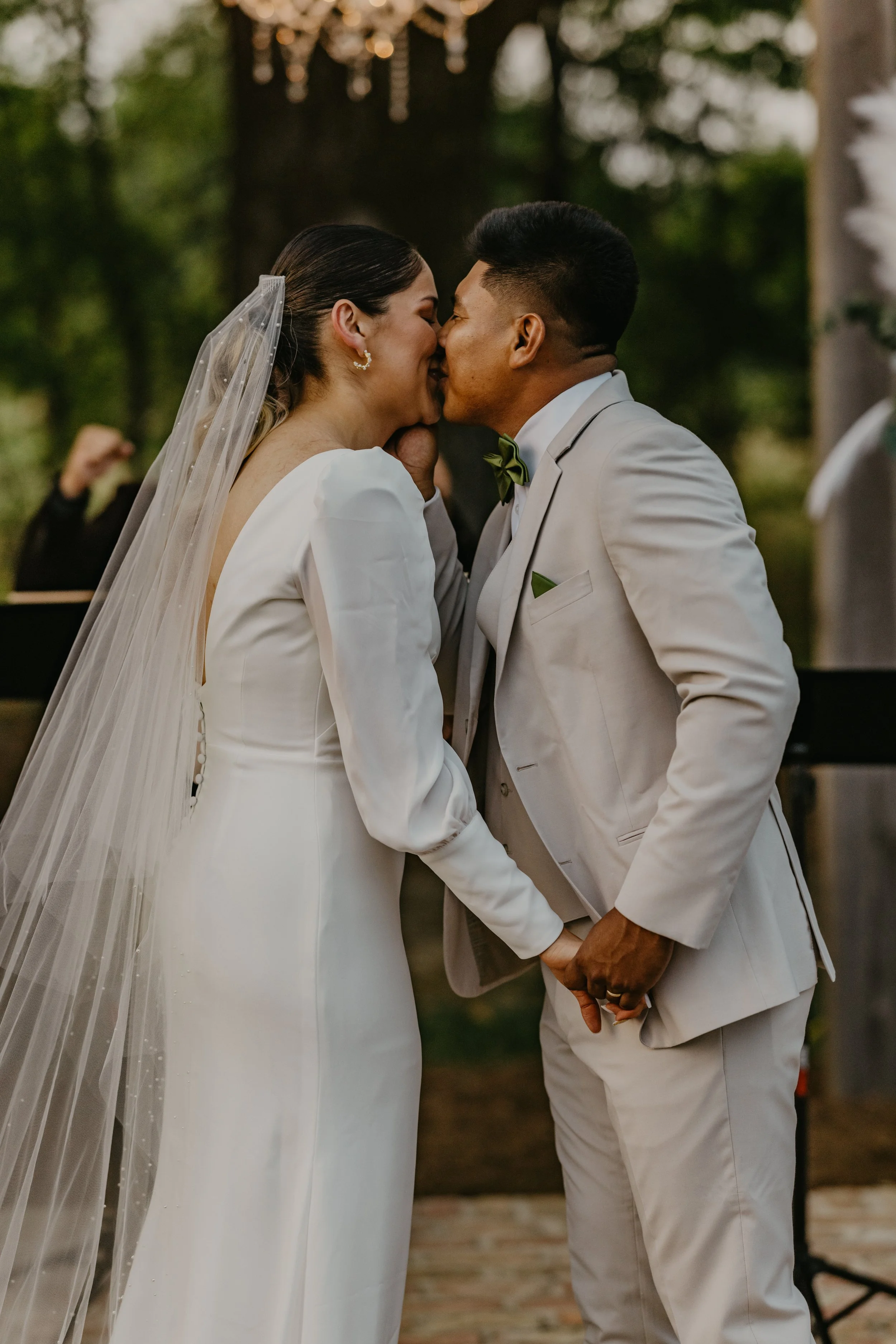A bride and groom share a kiss during their wedding ceremony outdoors. The bride is wearing a white gown with a veil, and the groom is in a light-colored suit with a green bow tie. They are holding hands in front of a tree decorated with hanging ligh
