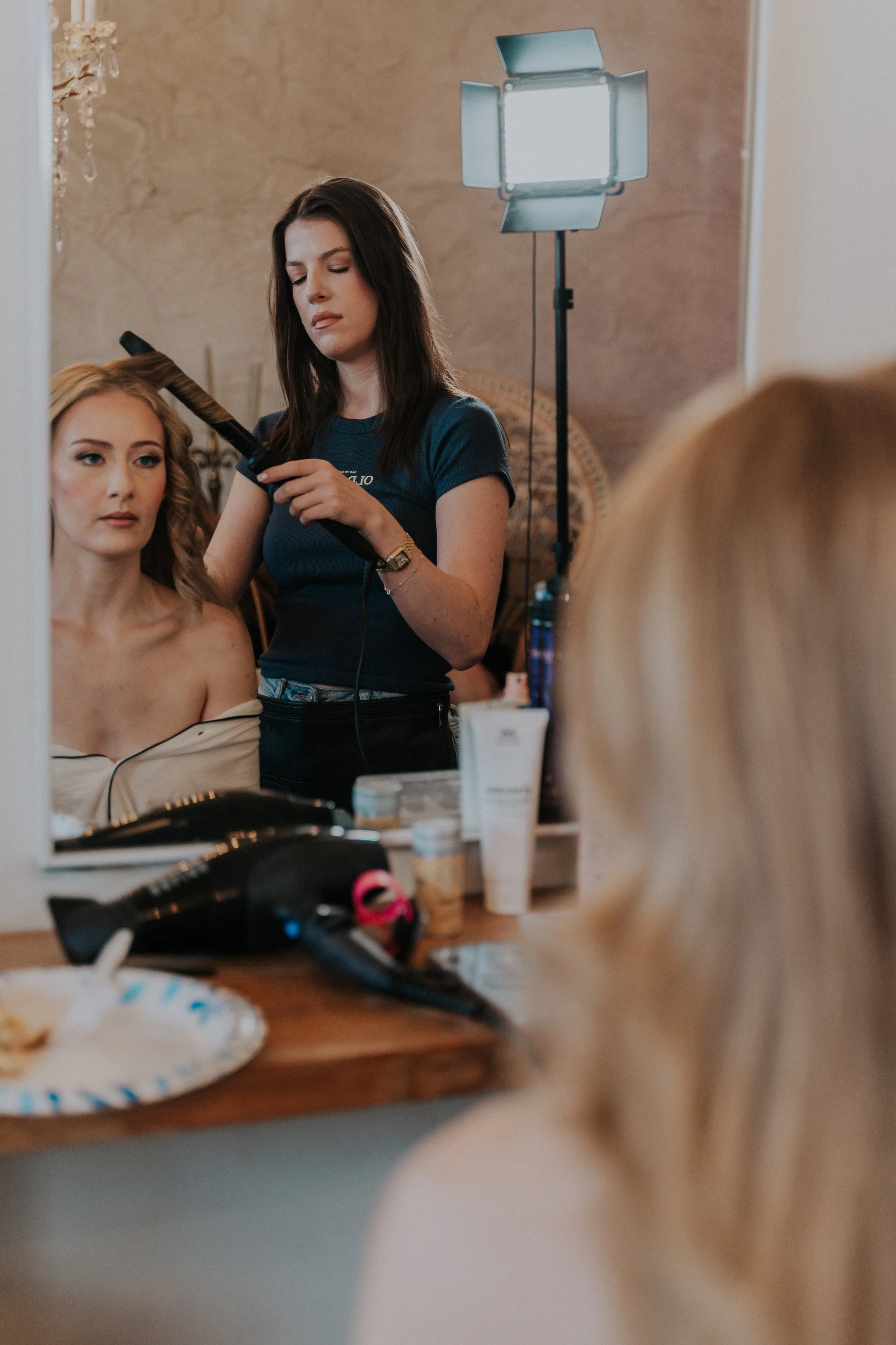 A makeup artist styles a woman with long wavy blonde hair sitting in front of a mirror, with a professional light setup and various beauty products on the table.