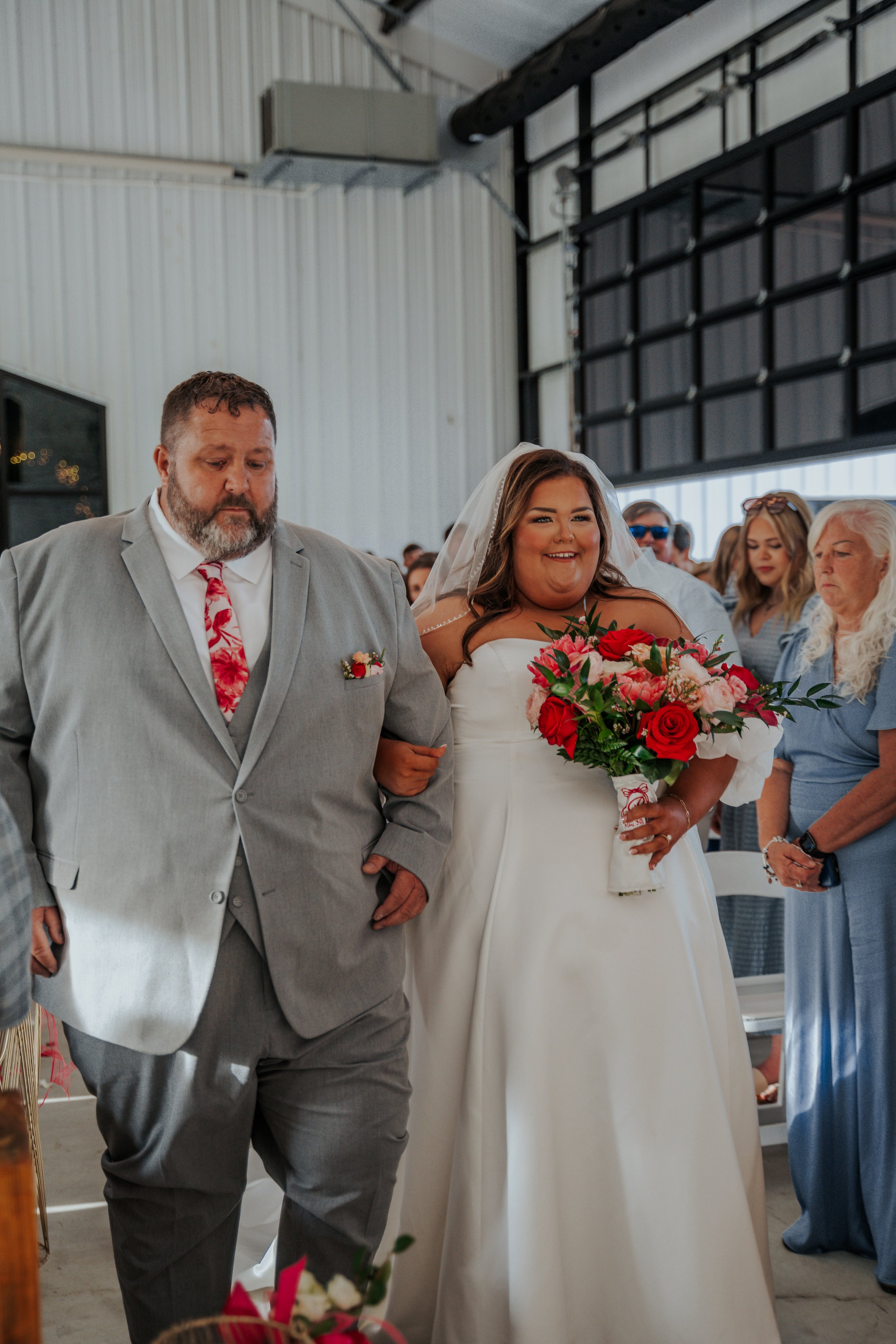 A bride in a white wedding gown holding a bouquet of red and pink flowers walking down the aisle, accompanied by a man in a gray suit, with guests watching.
