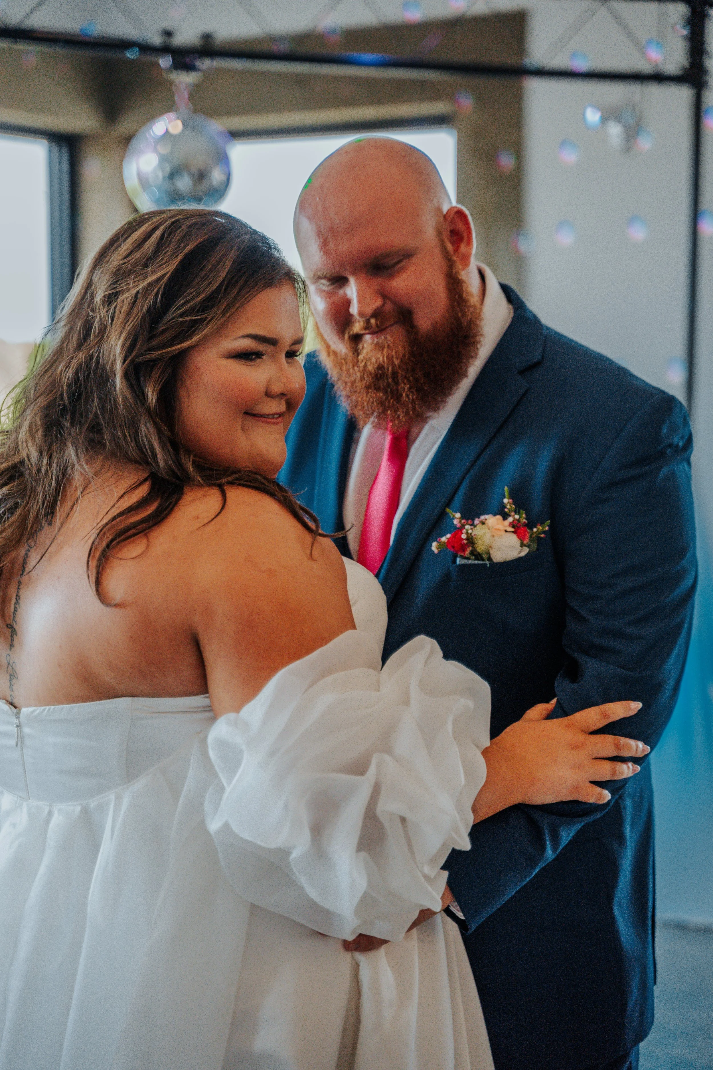 A bride and groom dancing at their wedding reception, smiling and looking at each other, with a disco ball and decorations in the background.