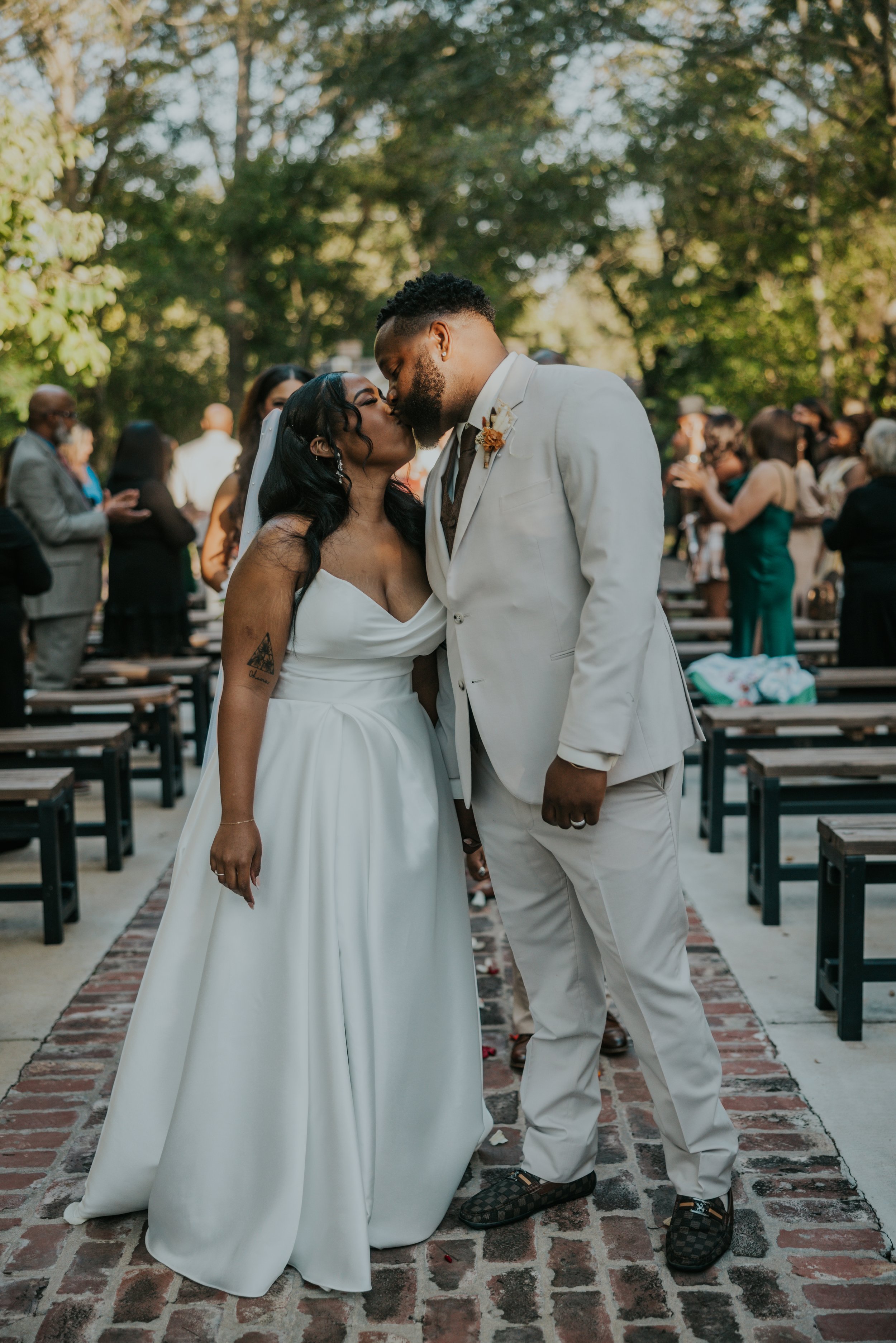 A bride and groom kiss during their outdoor wedding ceremony, with guests in the background and trees surrounding them.