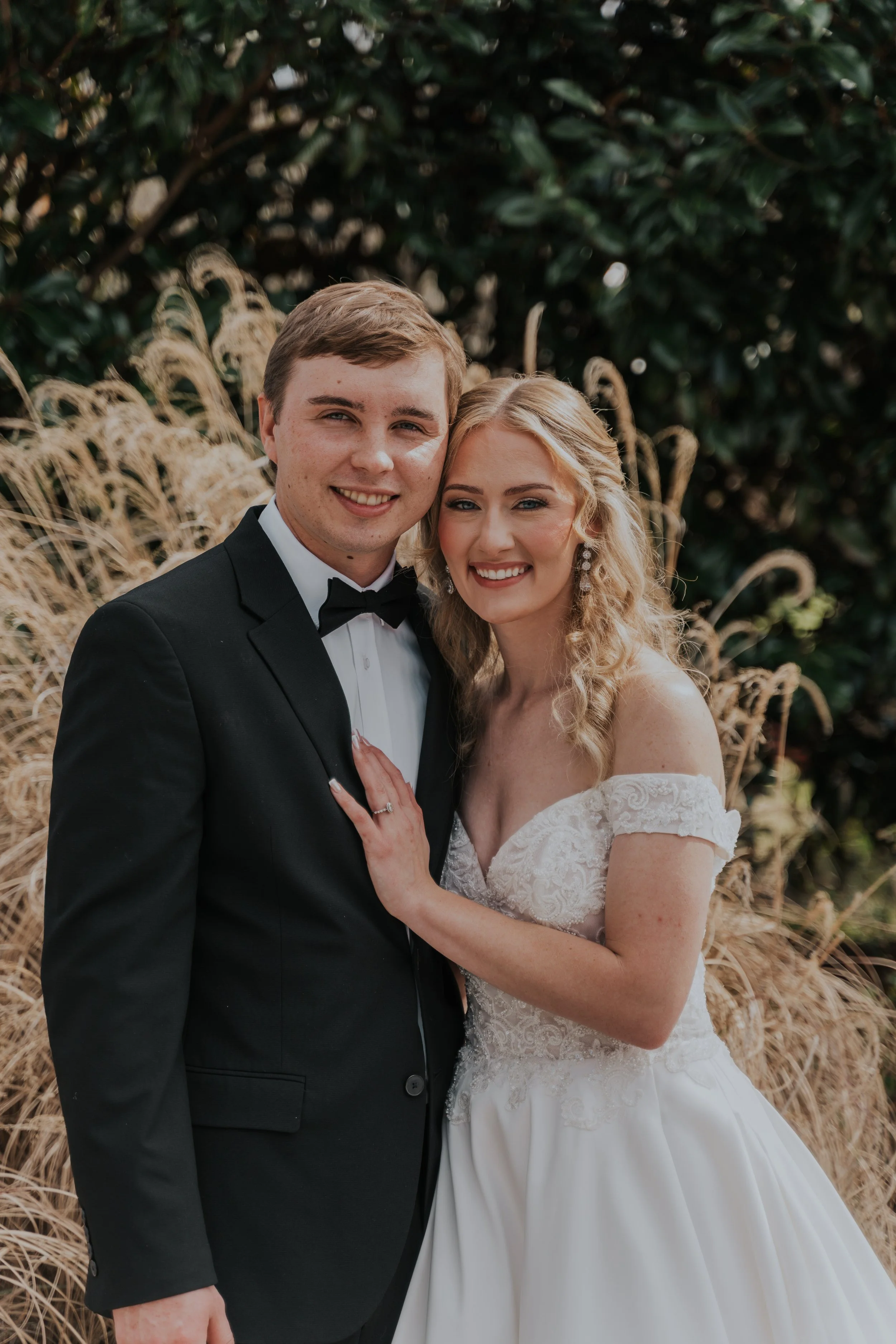 A newlywed couple in wedding attire, the groom in a black tuxedo and bow tie, the bride in a white lace off-the-shoulder wedding gown with curly blonde hair, standing outdoors with tall grasses and dark green foliage in the background.