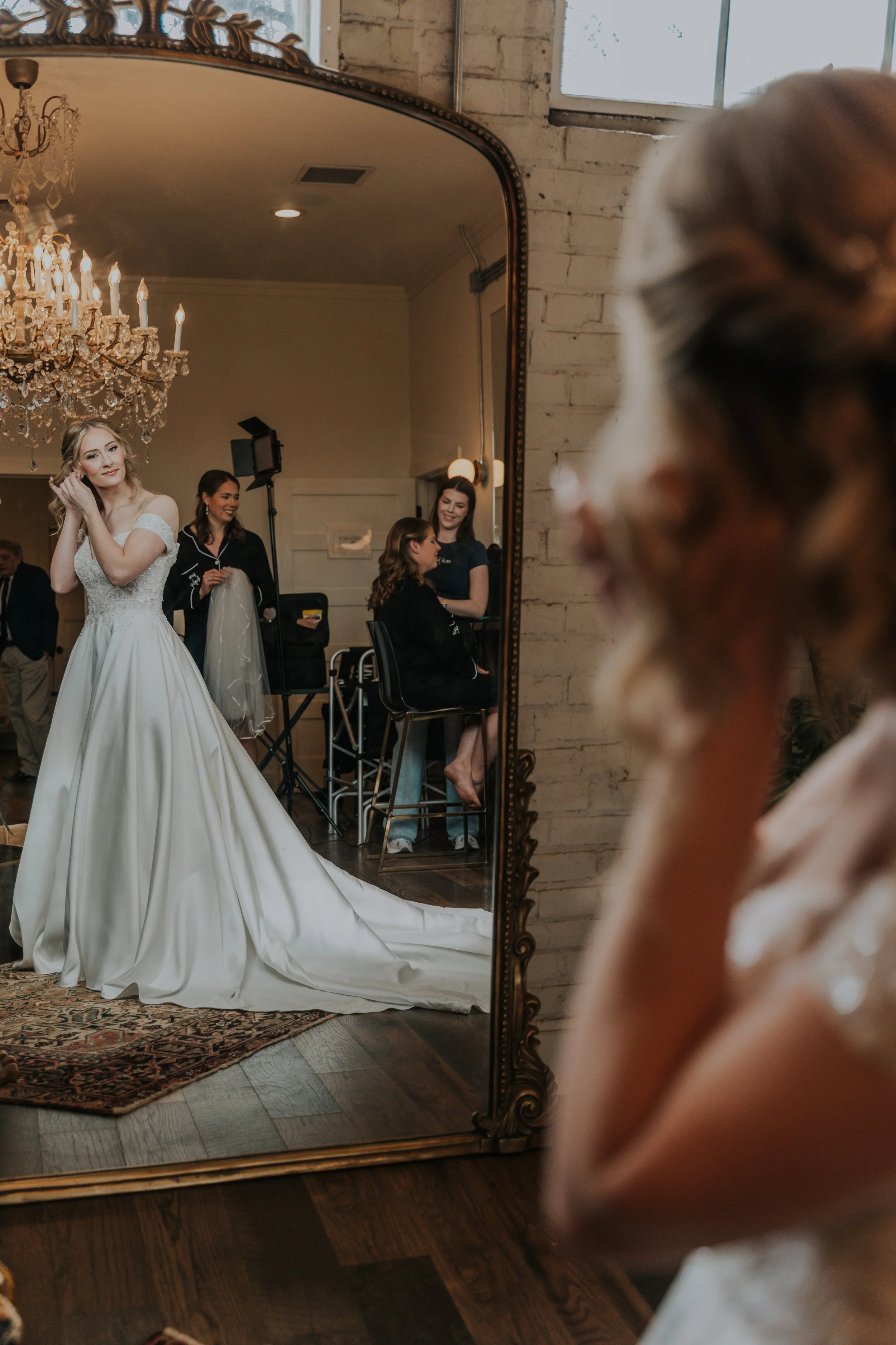A woman in a white wedding gown is looking into a mirror while getting ready, with her reflection showing her smiling and adjusting her hair. Two women and a girl are in the background, sitting and standing near the mirror, smiling and interacting.