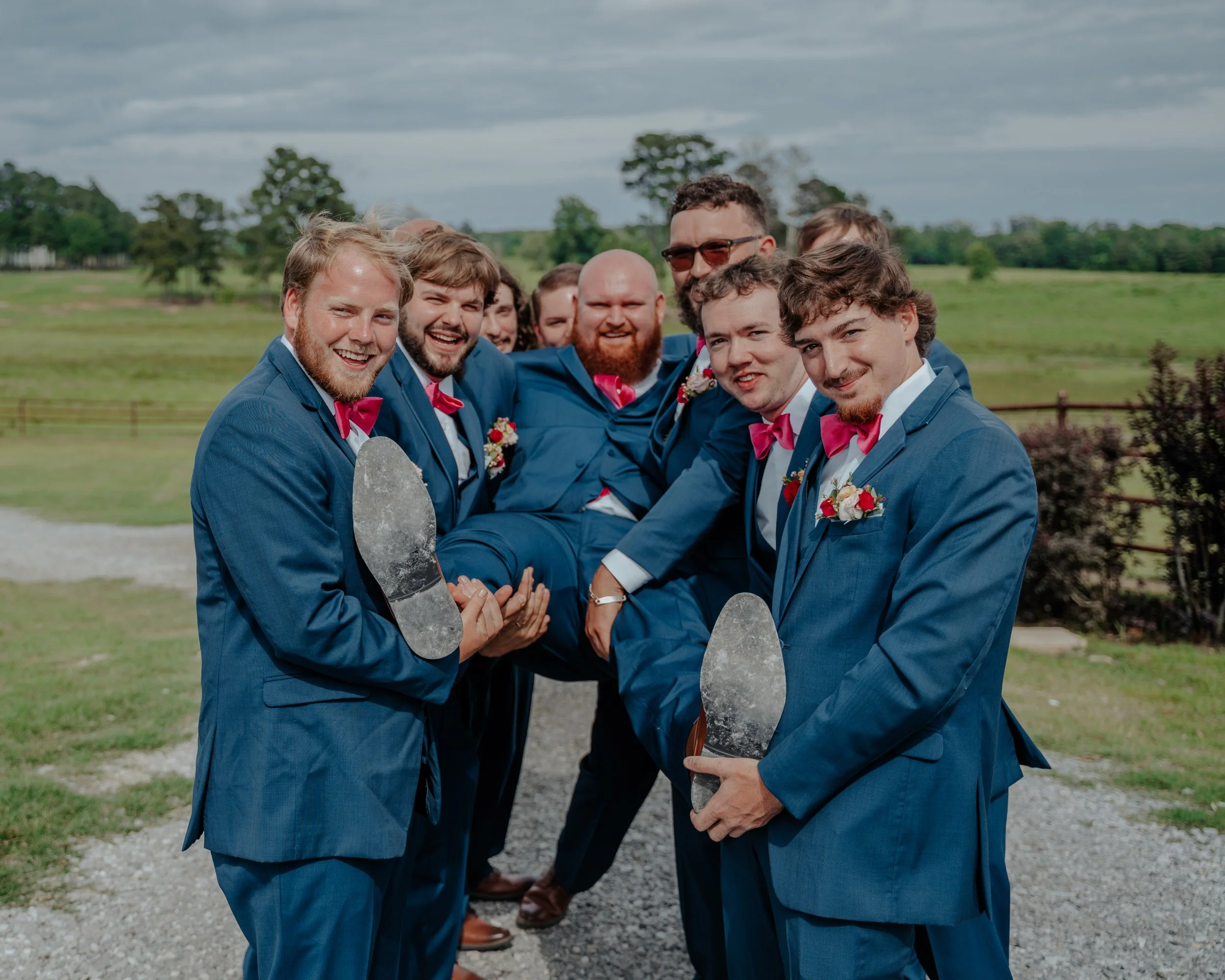 Group of men in blue suits with pink bow ties, some holding shoeprints, outdoor field, wedding celebration