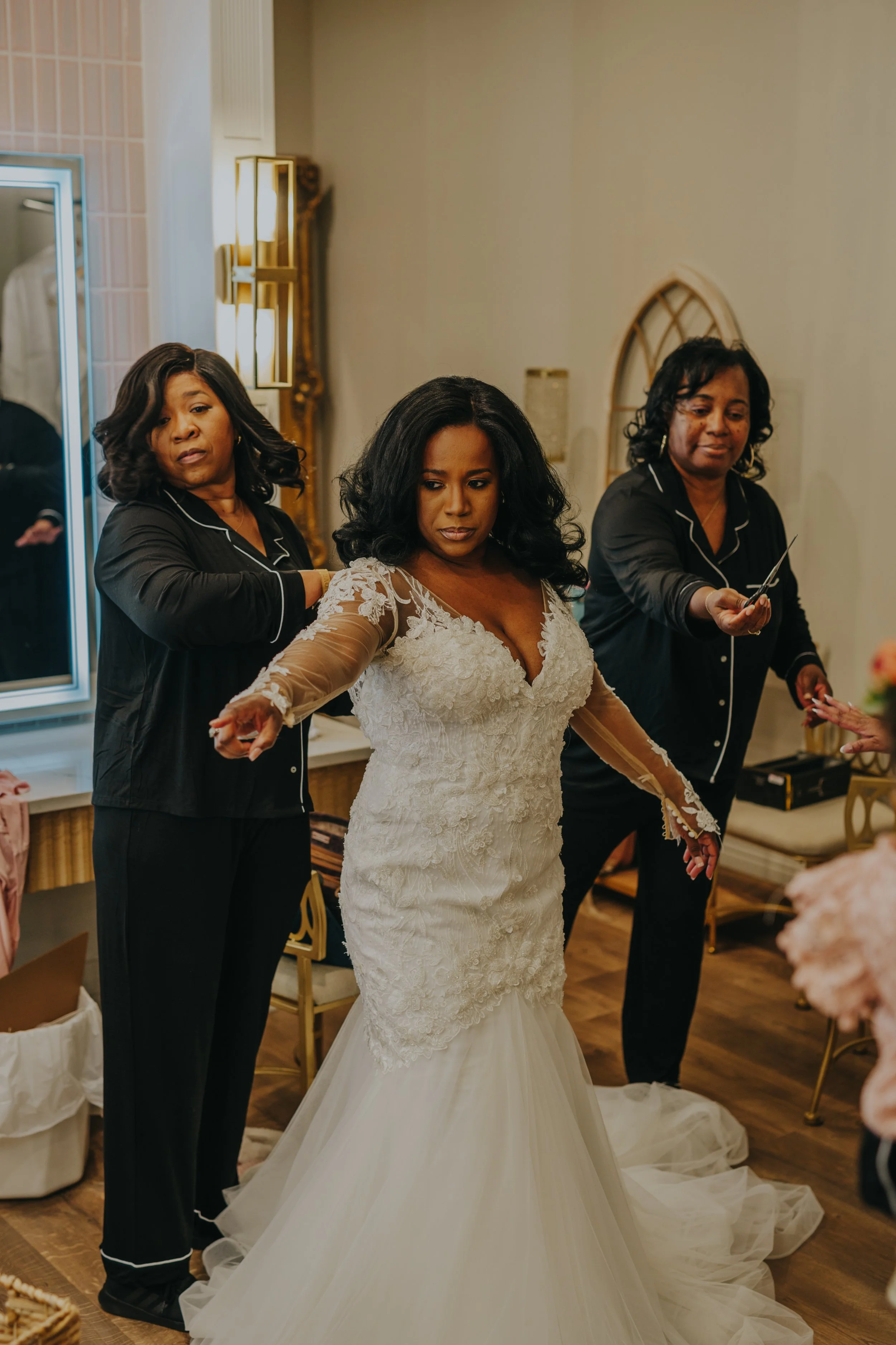 Bridal preparation as bride in lace wedding gown is assisted by two women in black pajamas.