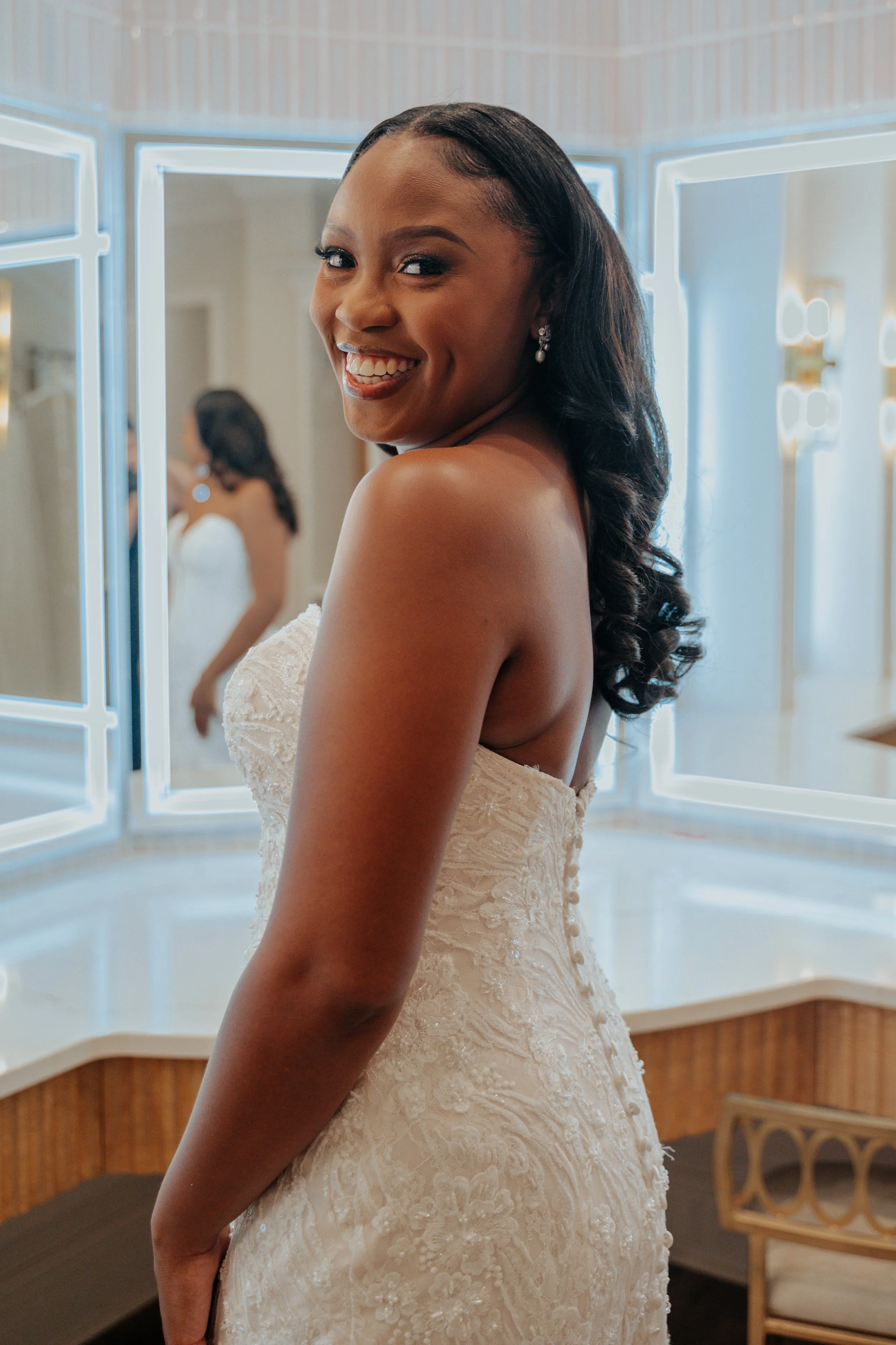 A woman in a wedding dress smiling at the camera, with a mirror and bright lights in the background.