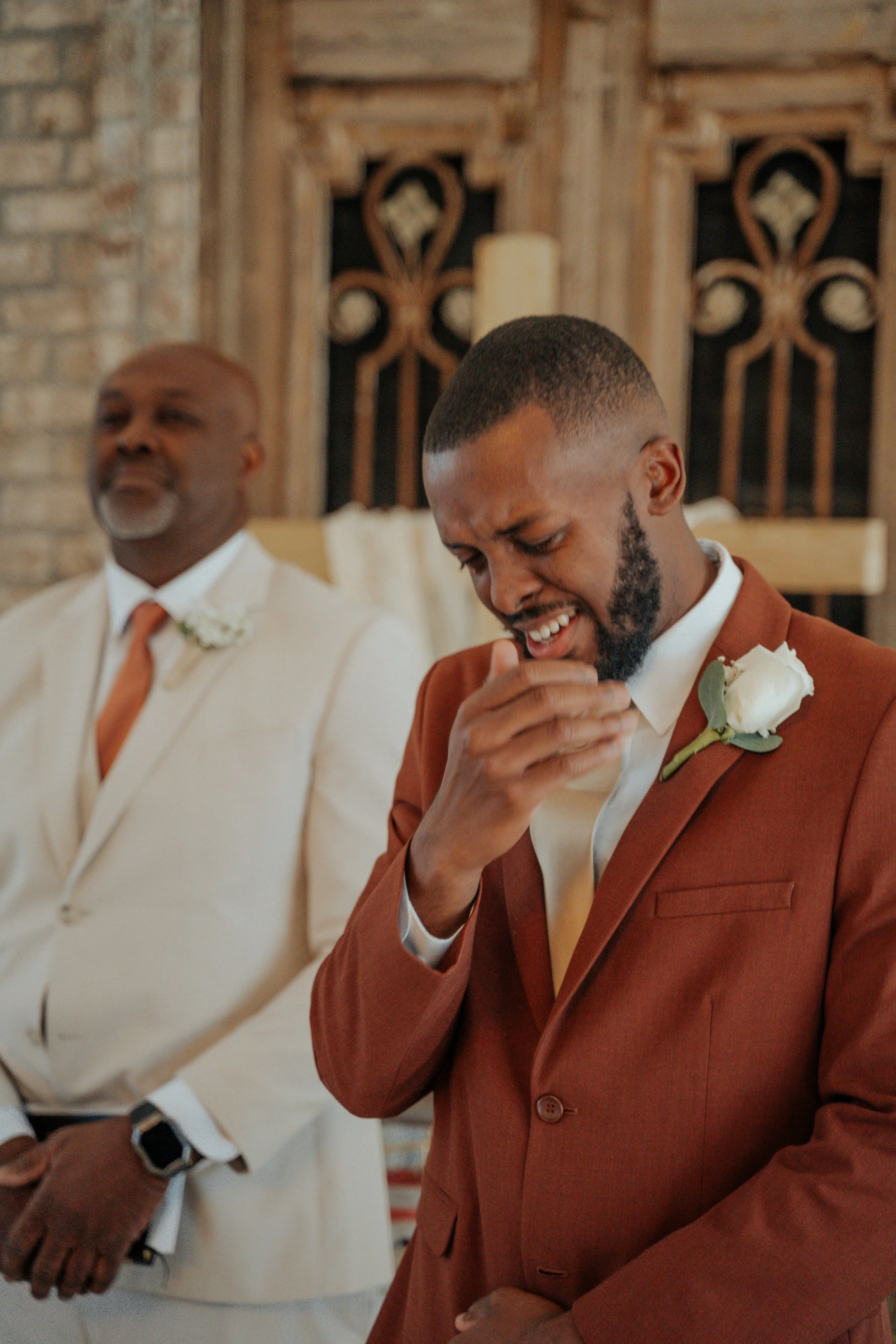 A groom in a rust-colored suit and white shirt, with a white rose boutonniere, appears emotional during a wedding ceremony, with an older man in a cream suit and white shirt standing beside him in a rustic setting.