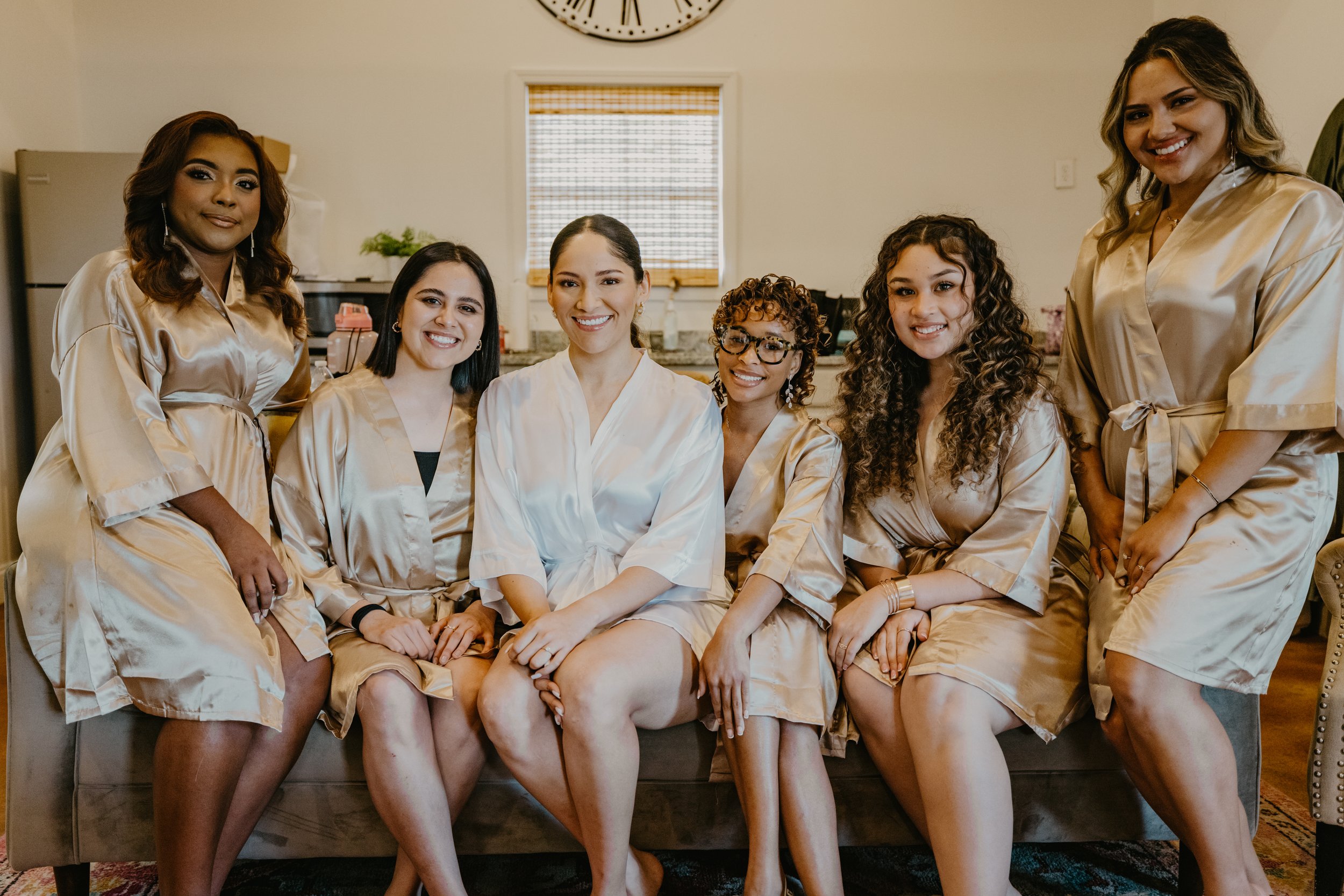 Six women sitting on a couch in matching gold satin robes, smiling in a kitchen with a window and a clock on the wall.