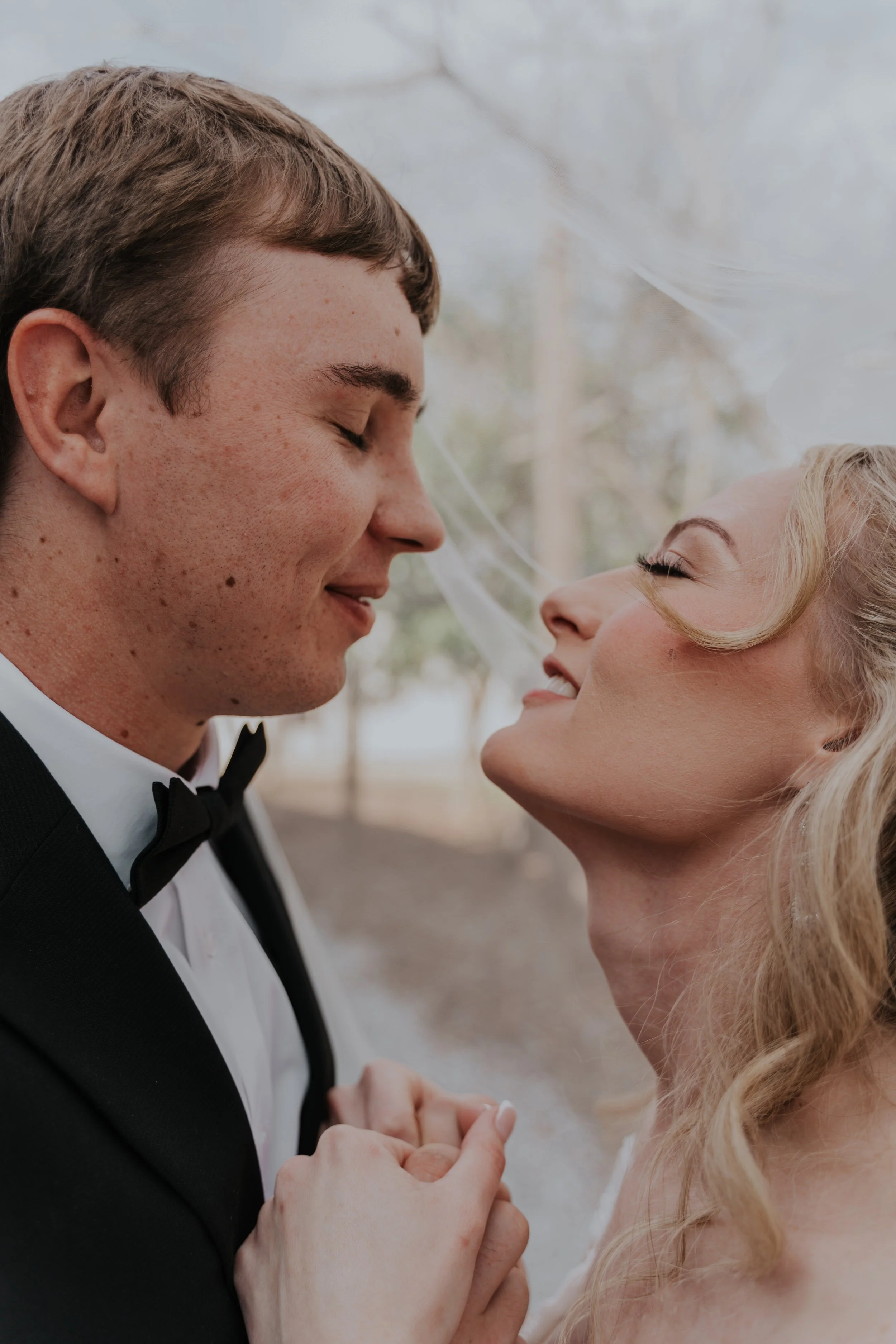 A newlywed couple, dressed in wedding attire, standing close with their foreheads touching, holding hands and smiling outdoors with blurred trees in the background.