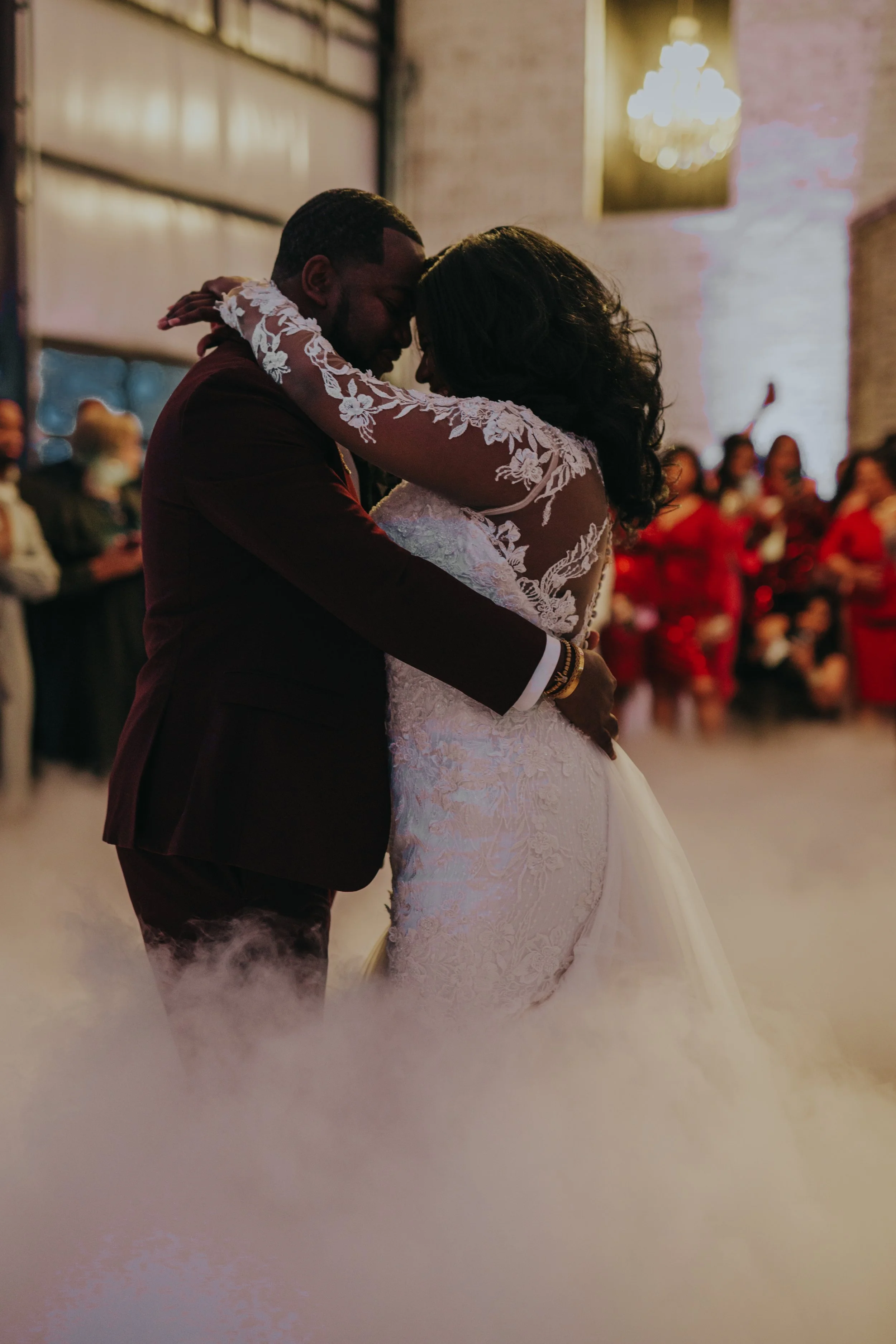 A couple in wedding attire dancing closely, with dark hair and skin, surrounded by an audience at a wedding reception. The man wears a dark suit, and the woman wears a white lace gown with floral embroidery.