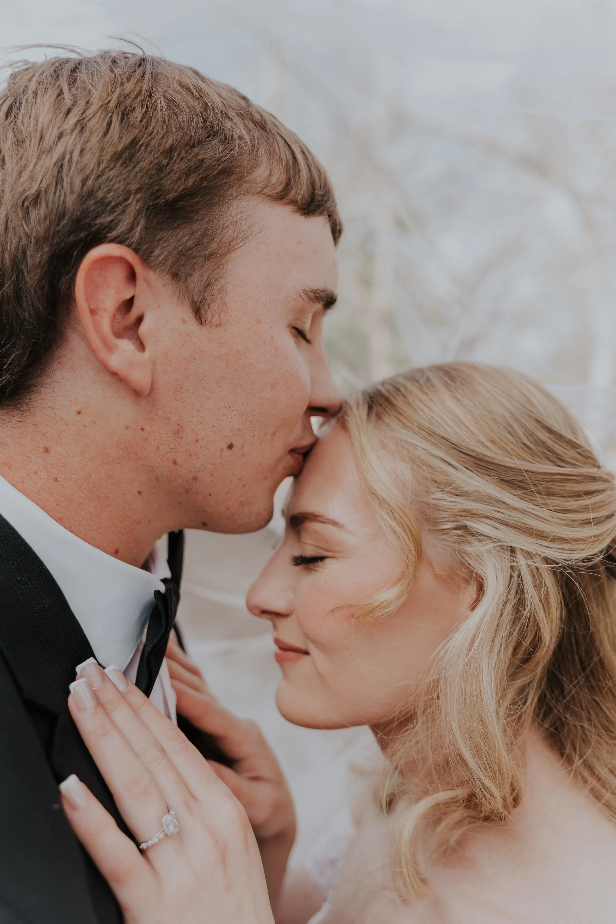 A close-up of a couple on their wedding day, with the groom gently pressing his forehead against the bride's, both with their eyes closed and serene expressions. The bride has blonde hair styled in loose waves and is wearing a wedding ring, while the