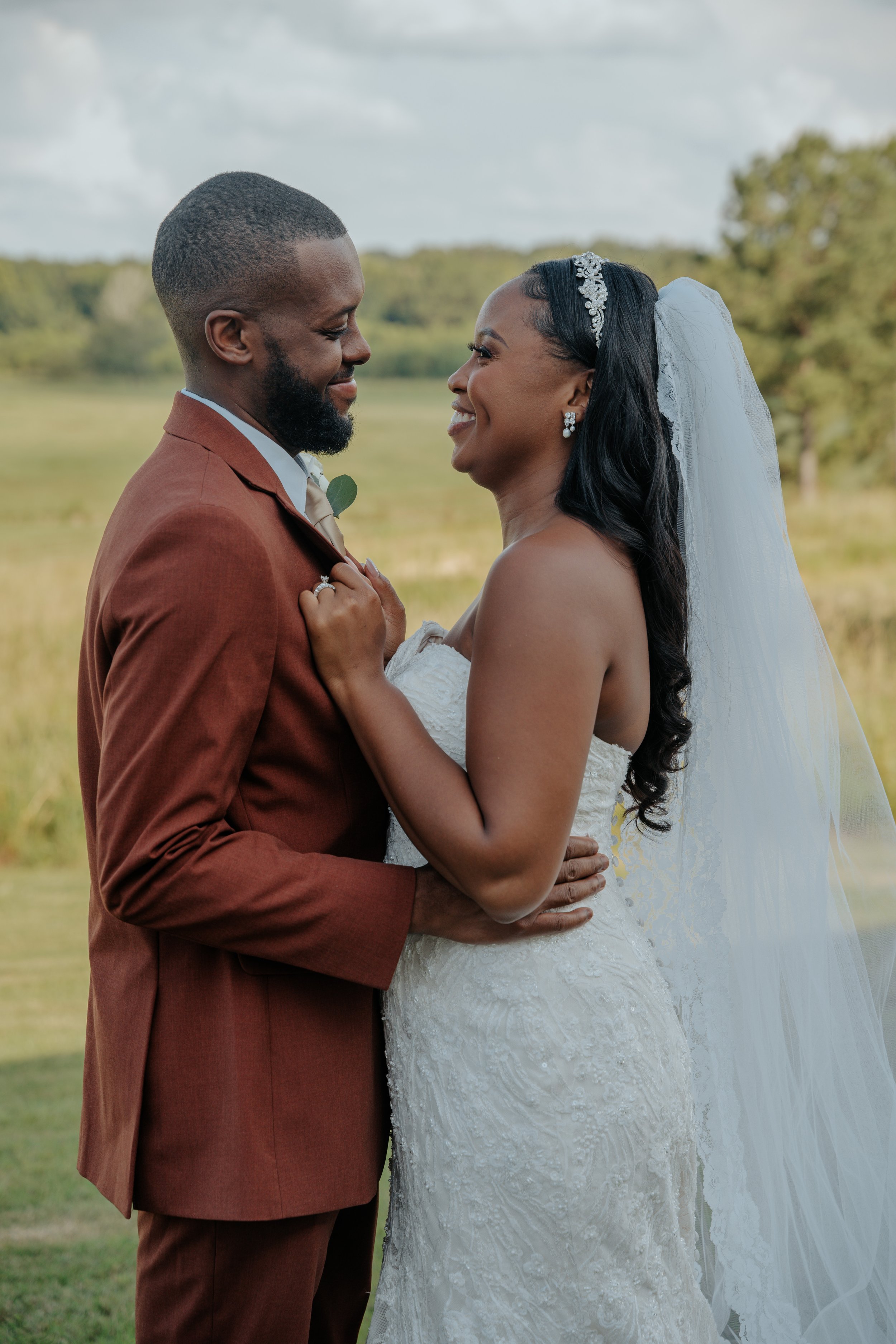 A bride and groom stand closely together outdoors, smiling at each other during a wedding ceremony.