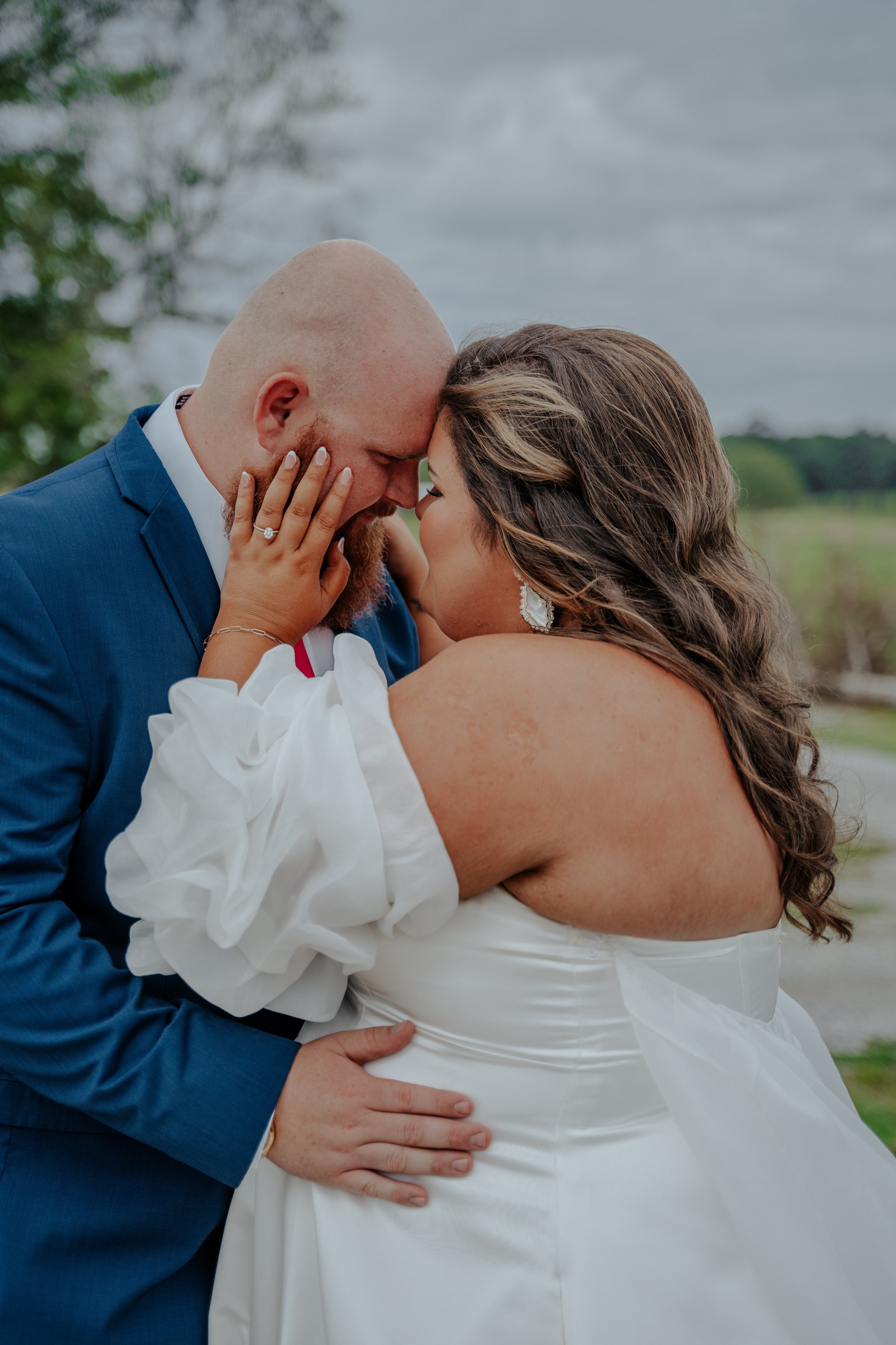 A couple is embracing outdoors, touching foreheads with their eyes closed, dressed in wedding attire.