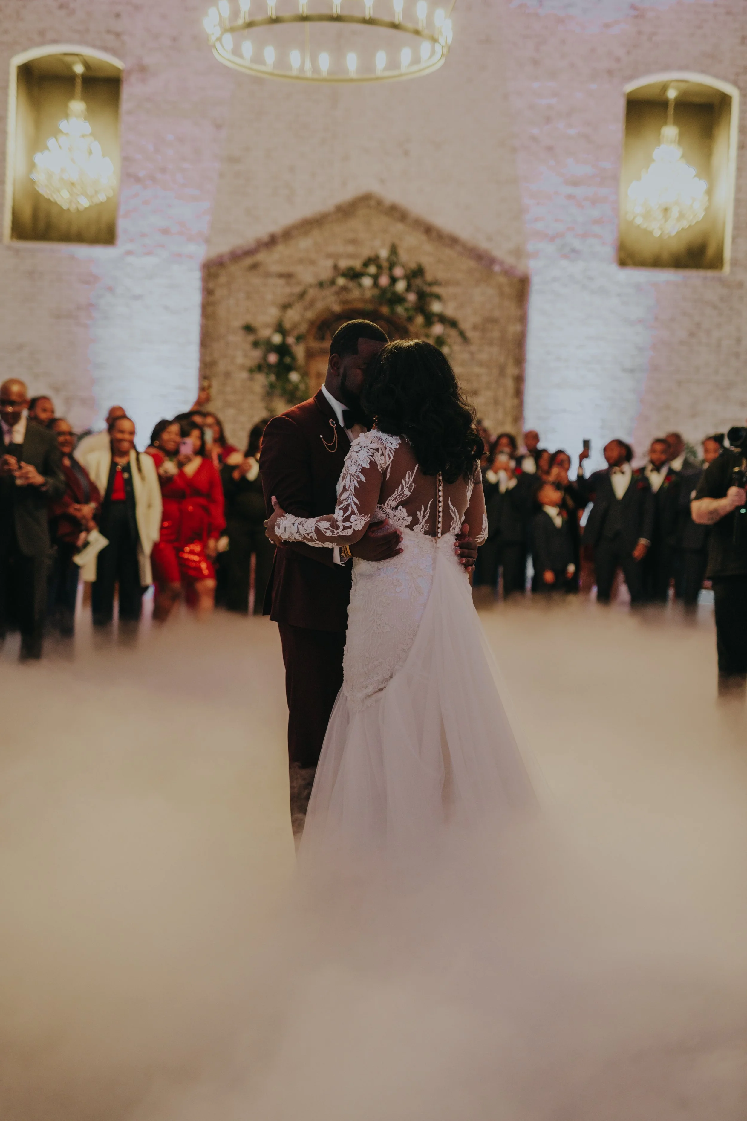 A couple is dancing during their wedding ceremony, surrounded by guests in a decorated venue with chandeliers and a brick wall background.