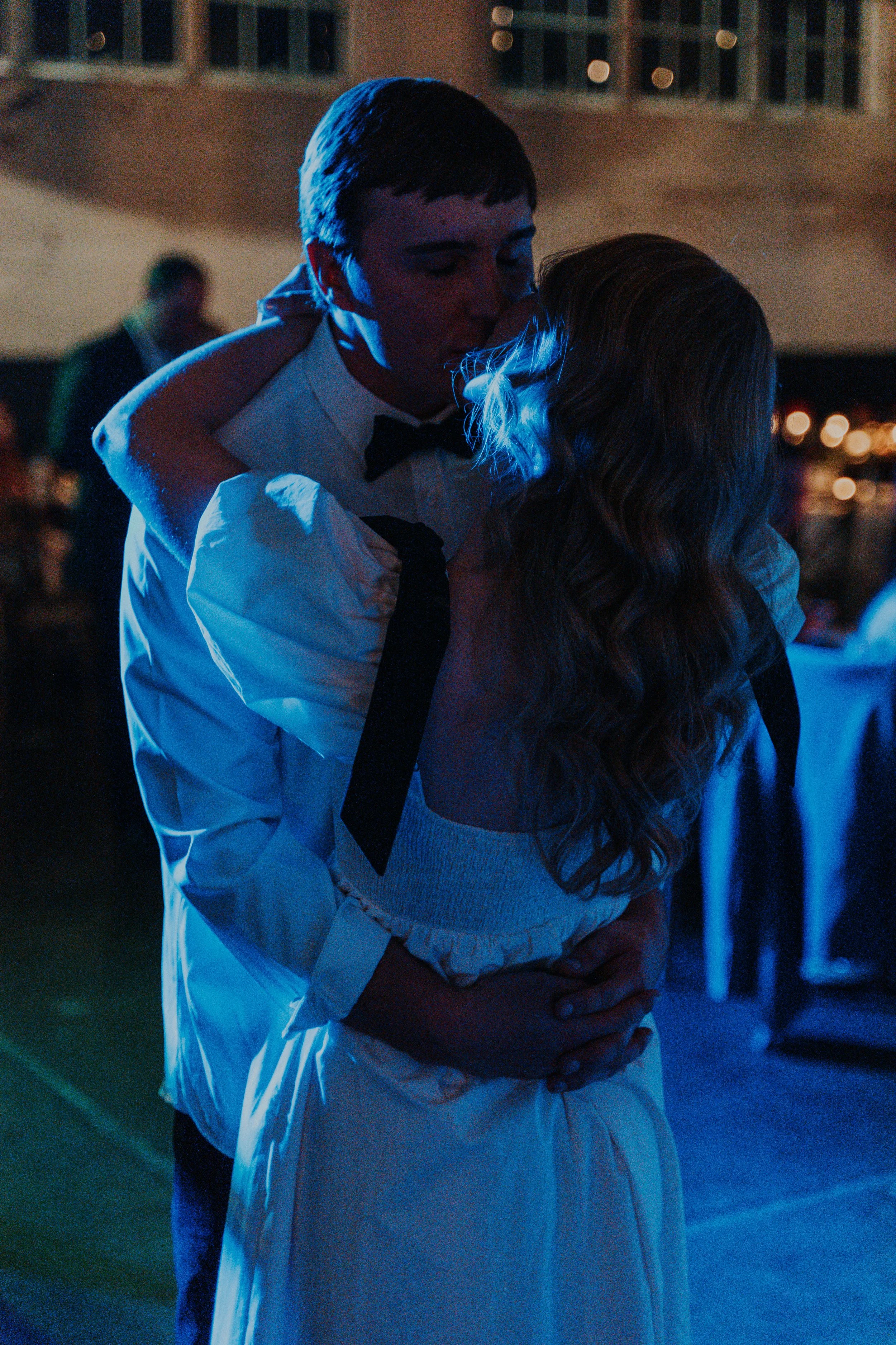 A young man in a tuxedo is kissing a woman in a white dress on the cheek at a wedding reception.