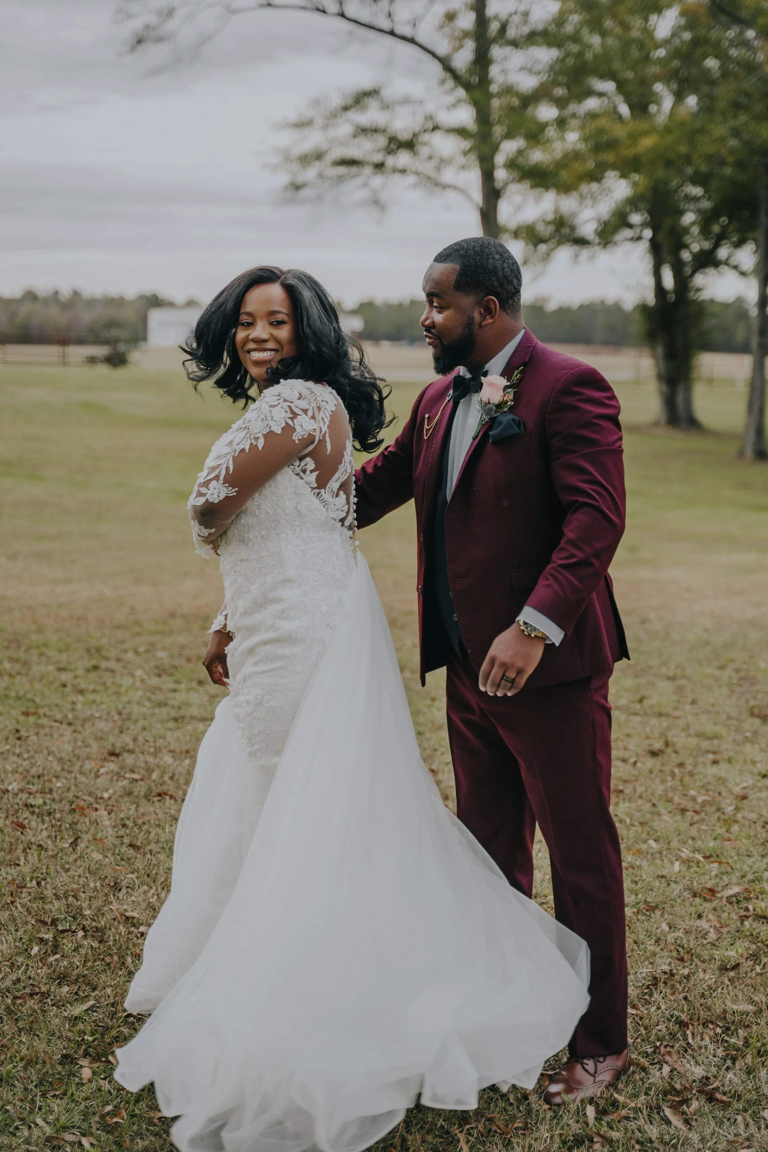 A bride and groom standing outdoors on a grassy field, dressed in wedding attire, with the bride smiling and the groom looking at her.