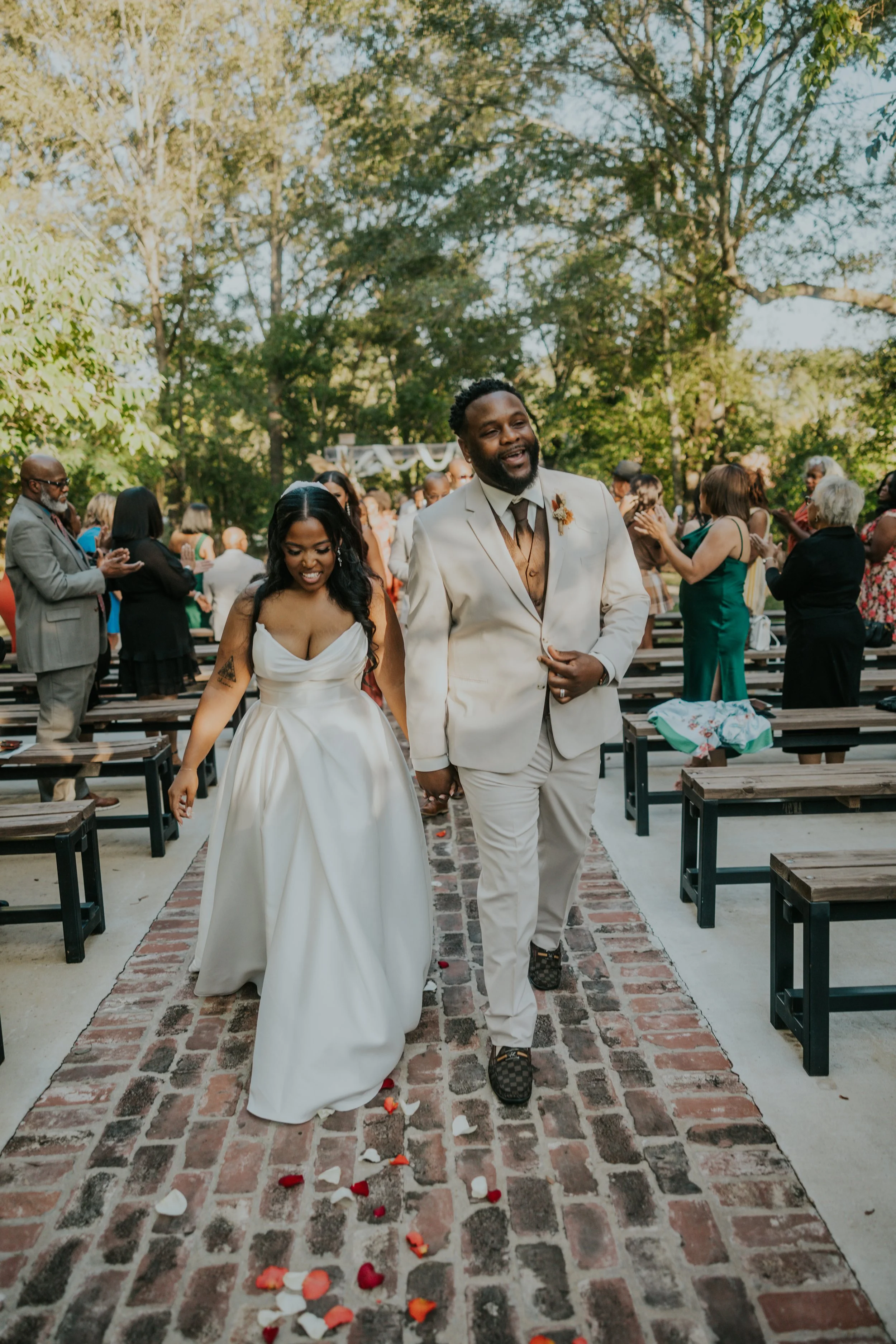 A bride and groom walking down the aisle at an outdoor wedding ceremony. The bride is wearing a white gown, and the groom is in a cream-colored suit. Guests are seated and clapping in the background.