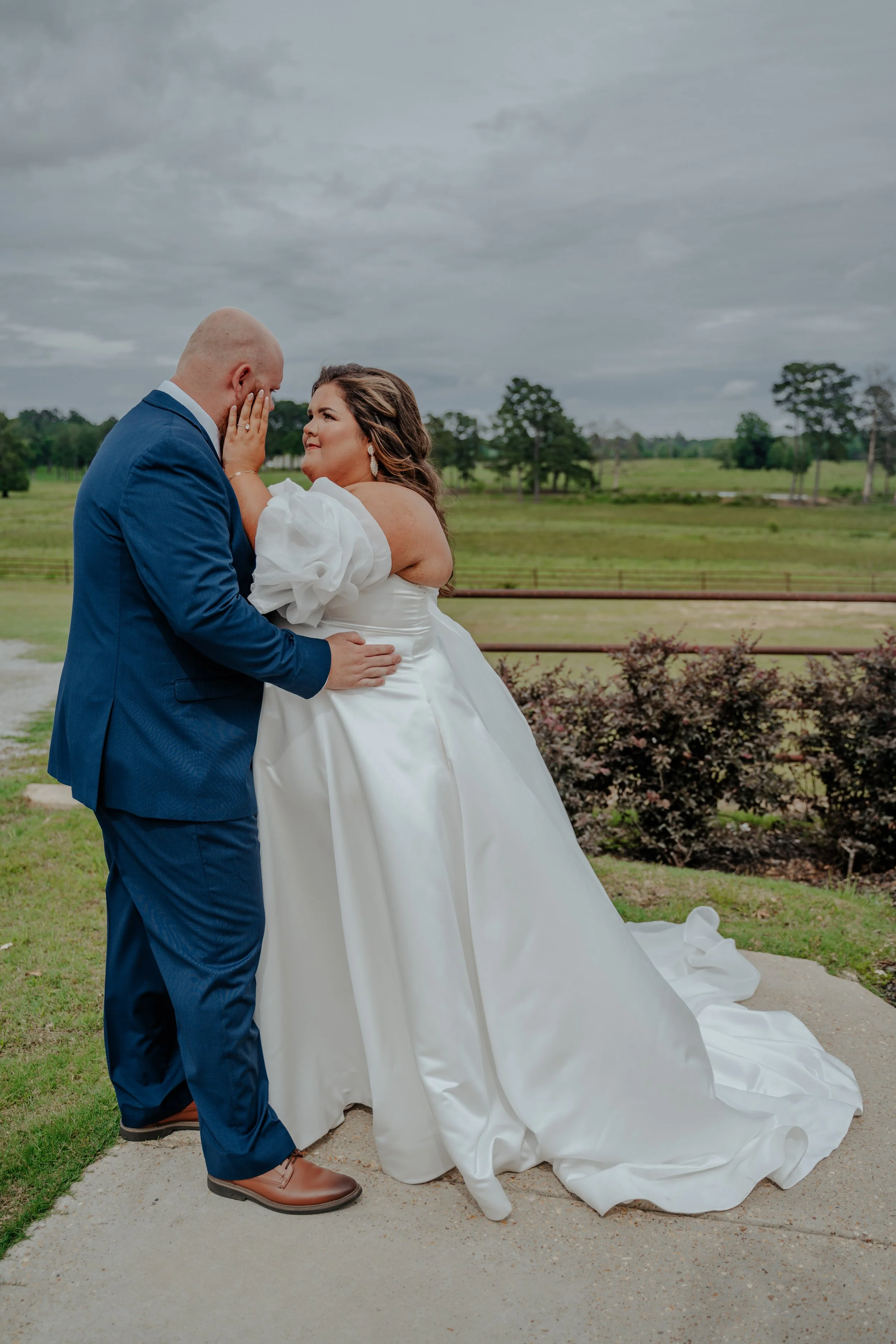 A bride and groom are sharing an emotional moment outdoors during a wedding, with the bride holding the groom's face and the groom holding her waist, against a cloudy sky and green field background.