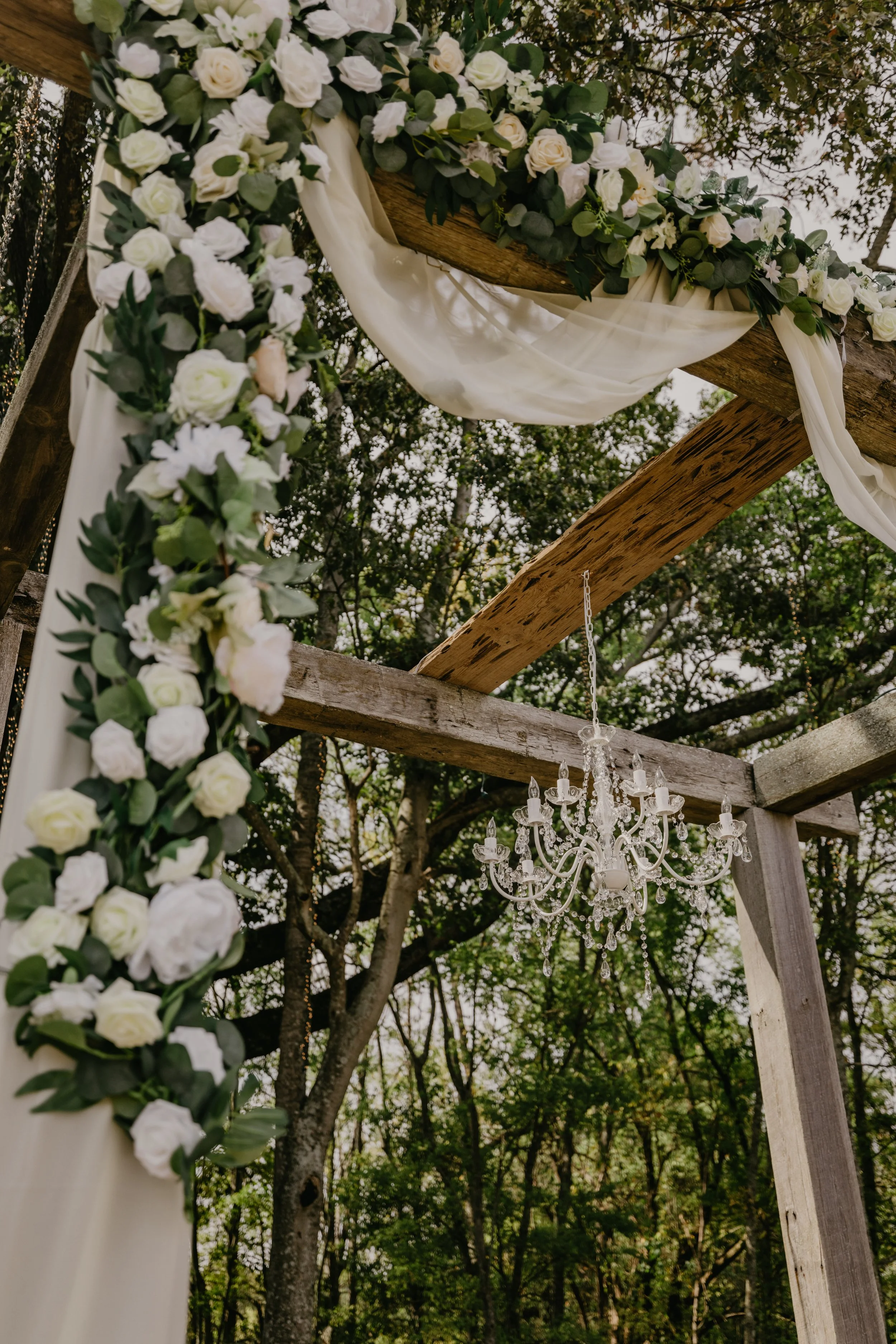 Floral wedding arch decorated with white roses, greenery, and sheer fabric, hanging under a chandelier in an outdoor setting with trees in the background.