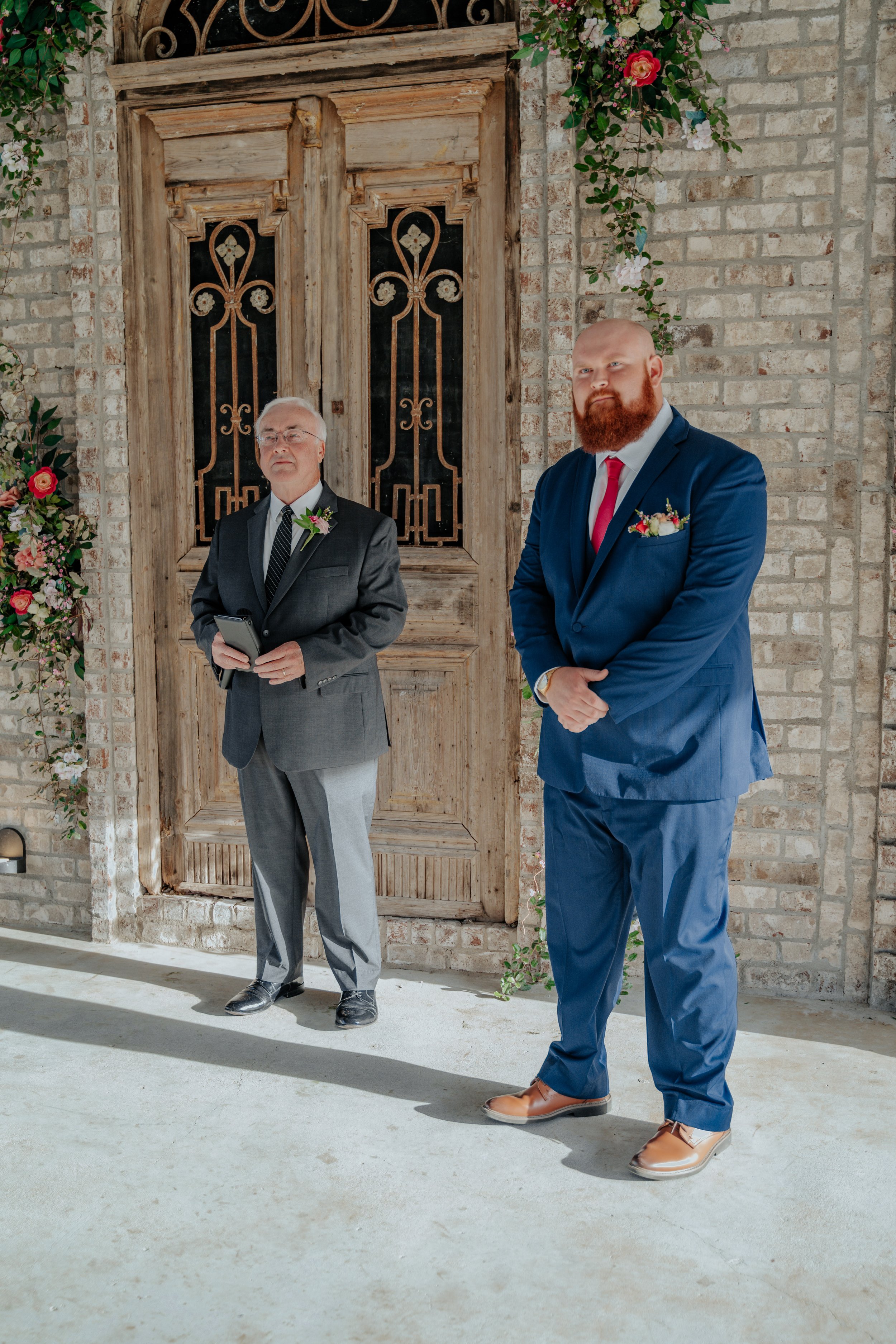 A groom in a blue suit and a man in a gray suit standing inside a rustic venue with brick walls and wooden door. The groom is smiling with his hands clasped, and the man is holding a phone. Floral decorations are around the doorway.