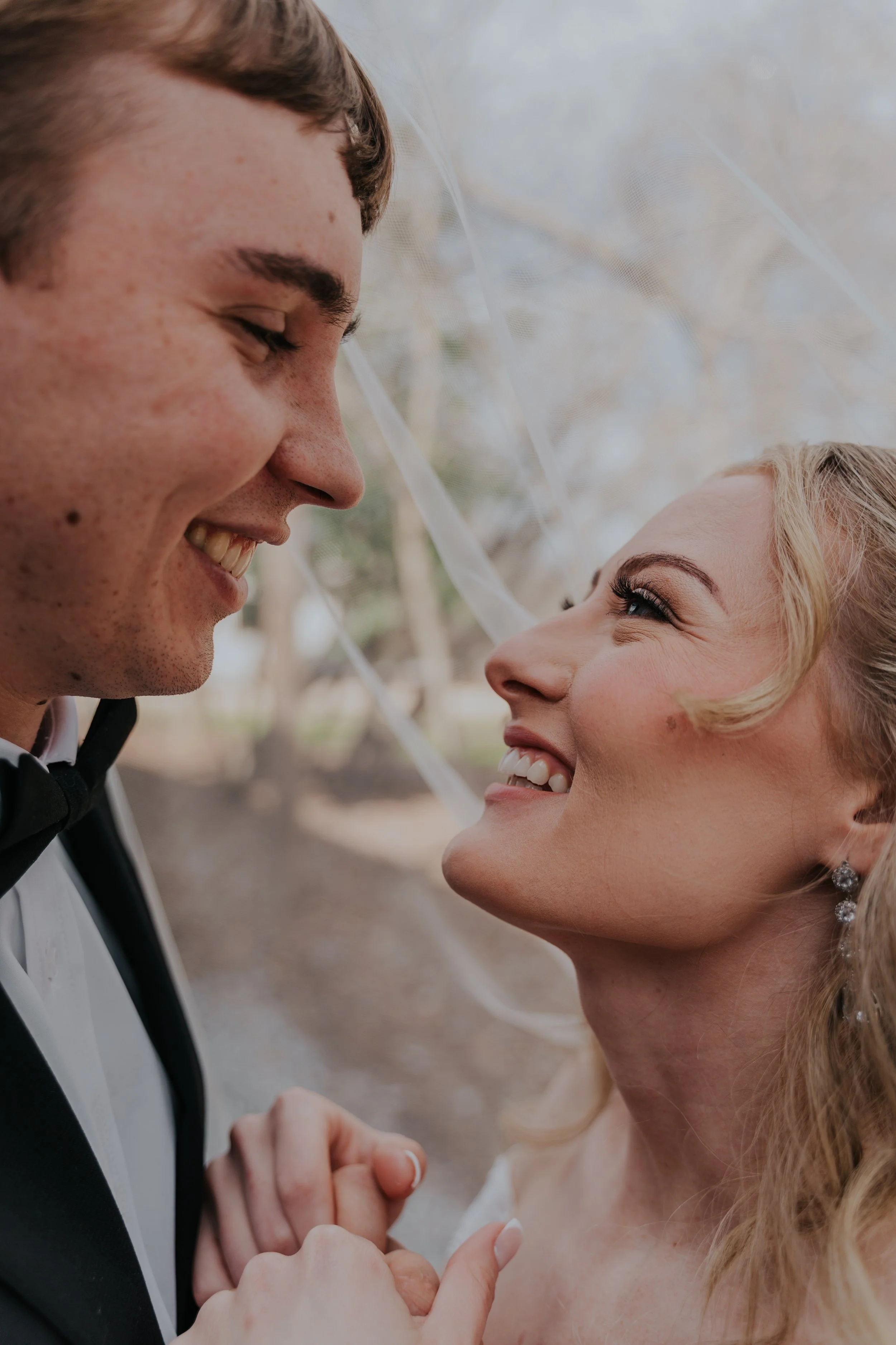 A bride and groom are smiling and looking into each other's eyes in an outdoor wedding setting.