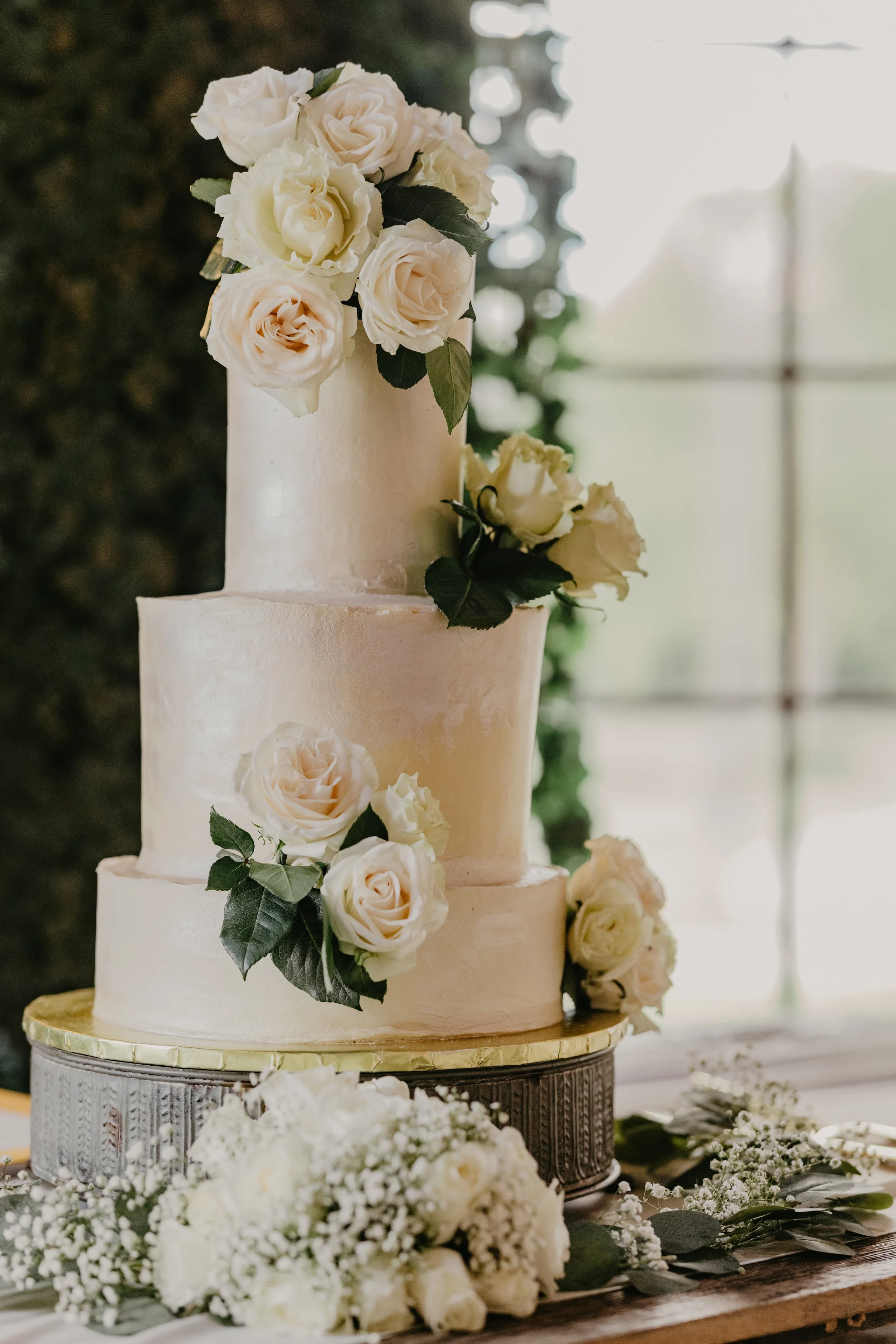 A three-tiered white wedding cake decorated with white roses and greenery, with floral arrangements at the base and on top, sitting on a decorative cake stand.