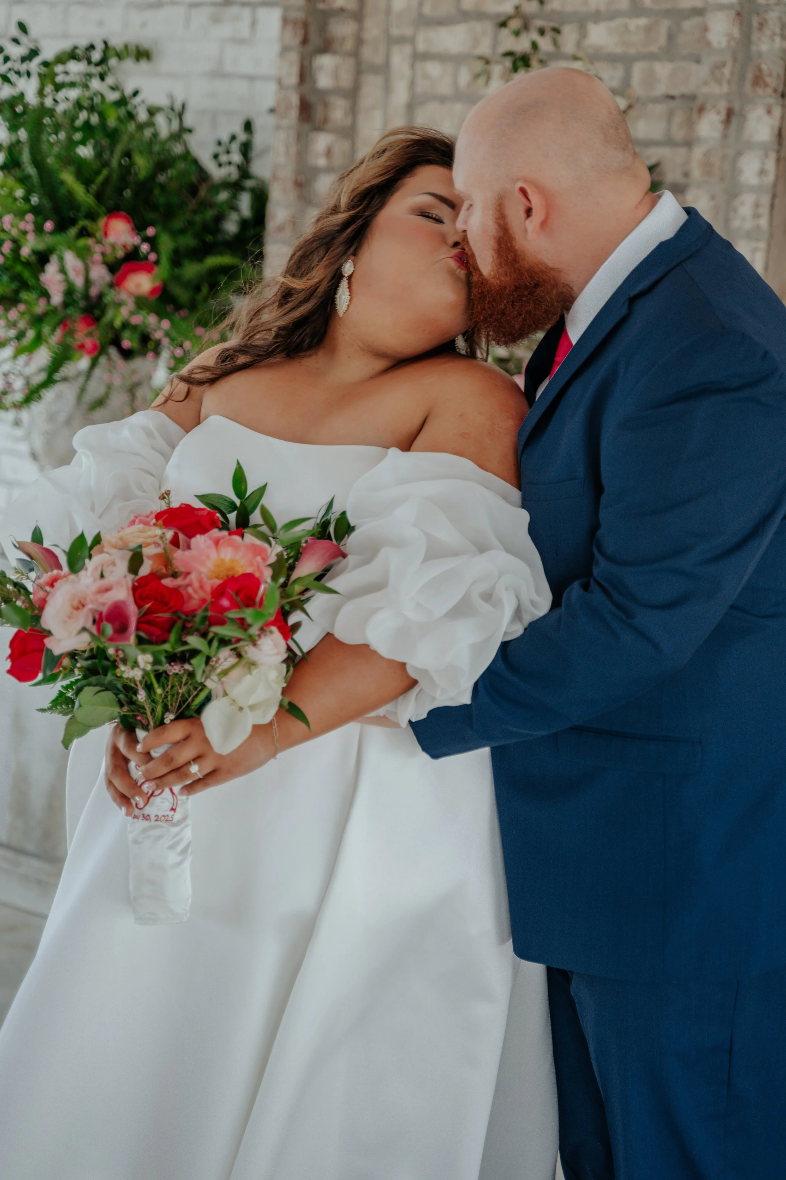 Bride and groom sharing a kiss during their wedding ceremony, with the bride holding a bouquet of pink, red, and white flowers, and background featuring floral arrangements and brick wall.