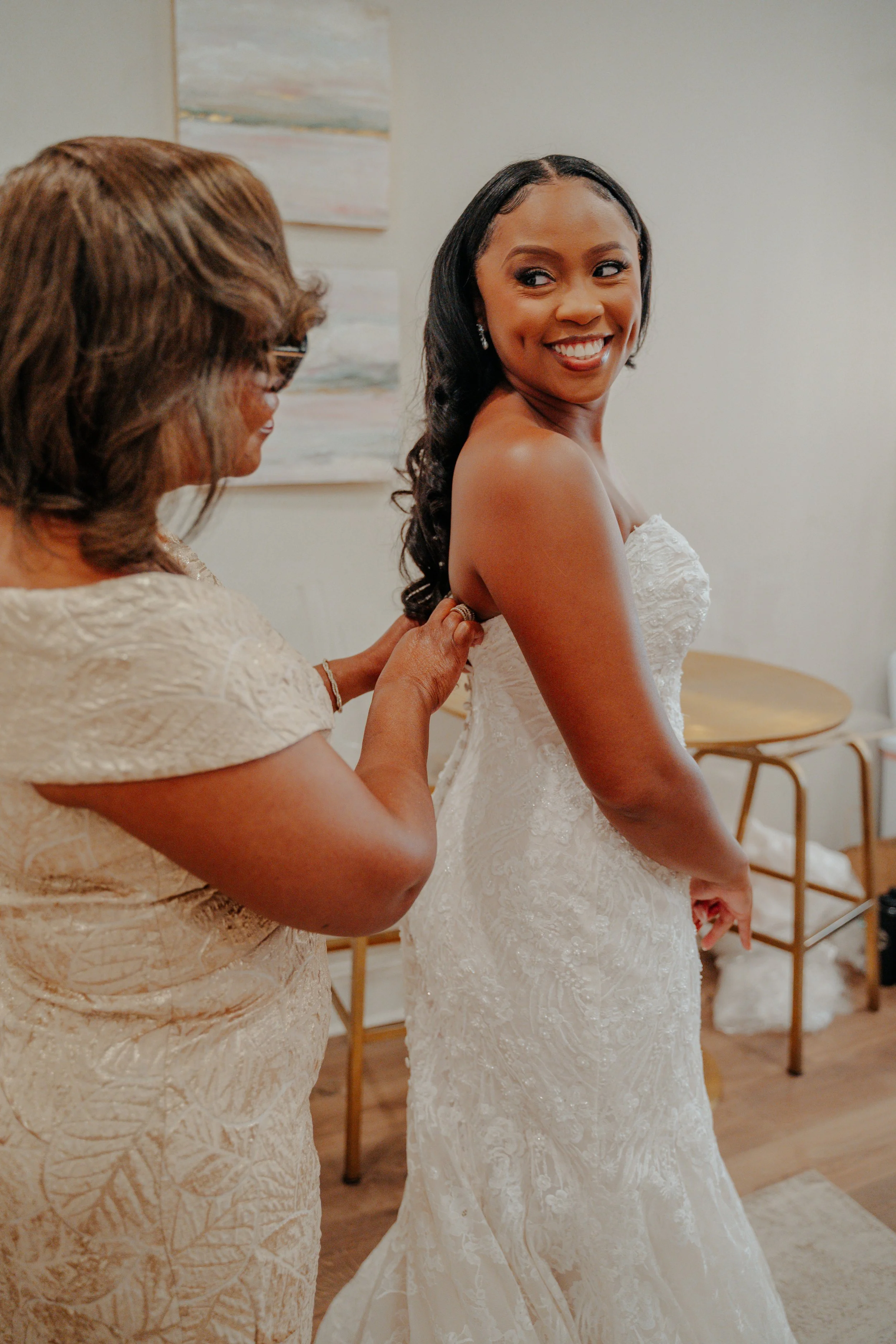 A bride in a white wedding dress getting ready for her wedding while her mother helps her zip up the back of her gown.
