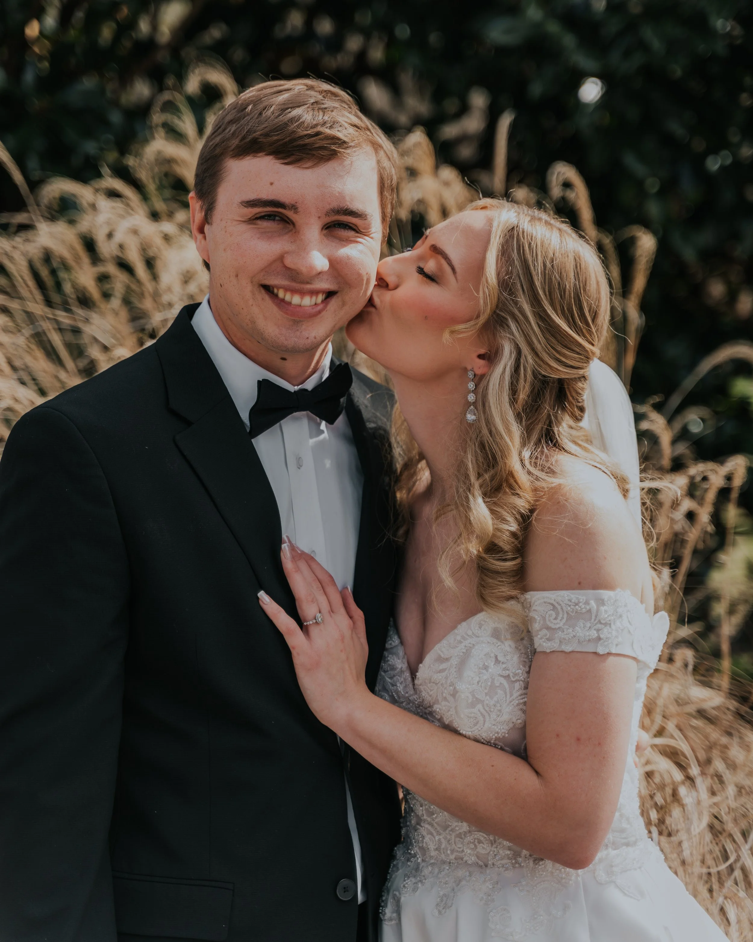 A newlywed couple, the groom in a black tuxedo and bowtie, and the bride in a white wedding dress with off-the-shoulder lace sleeves, sharing a kiss outside. The bride is kissing the groom on the cheek, and they are smiling at the camera, with tall g