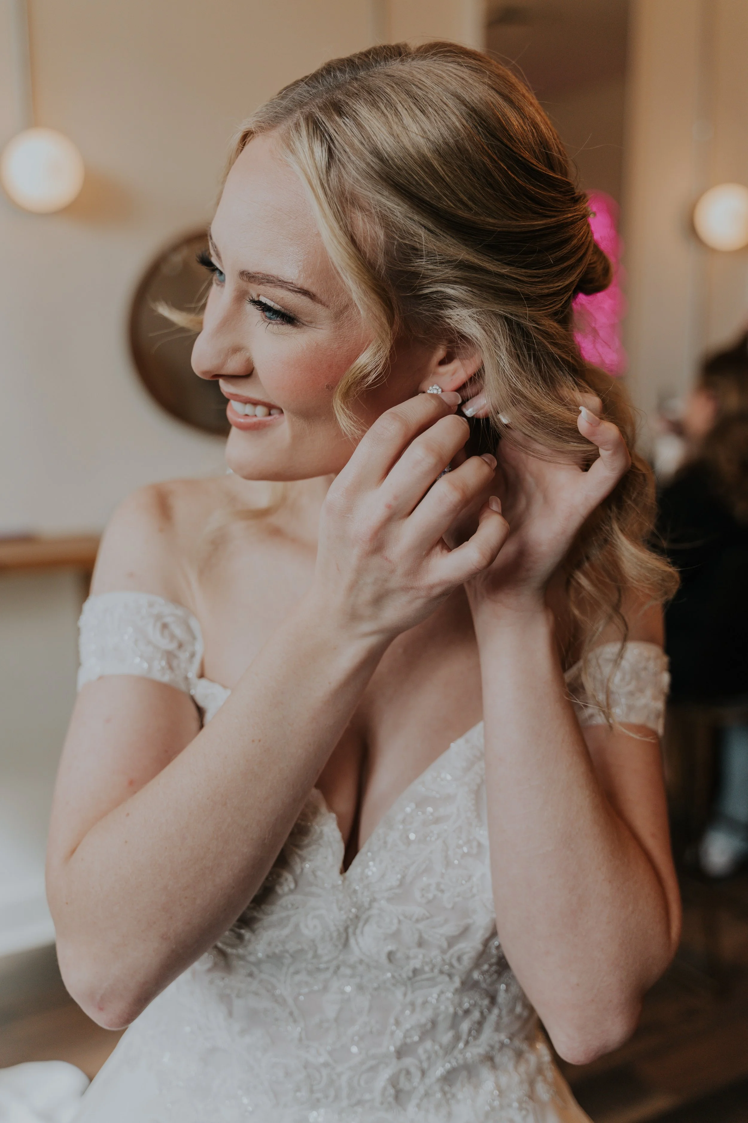 Bride putting on earrings before her wedding, smiling in a white off-shoulder lace dress.