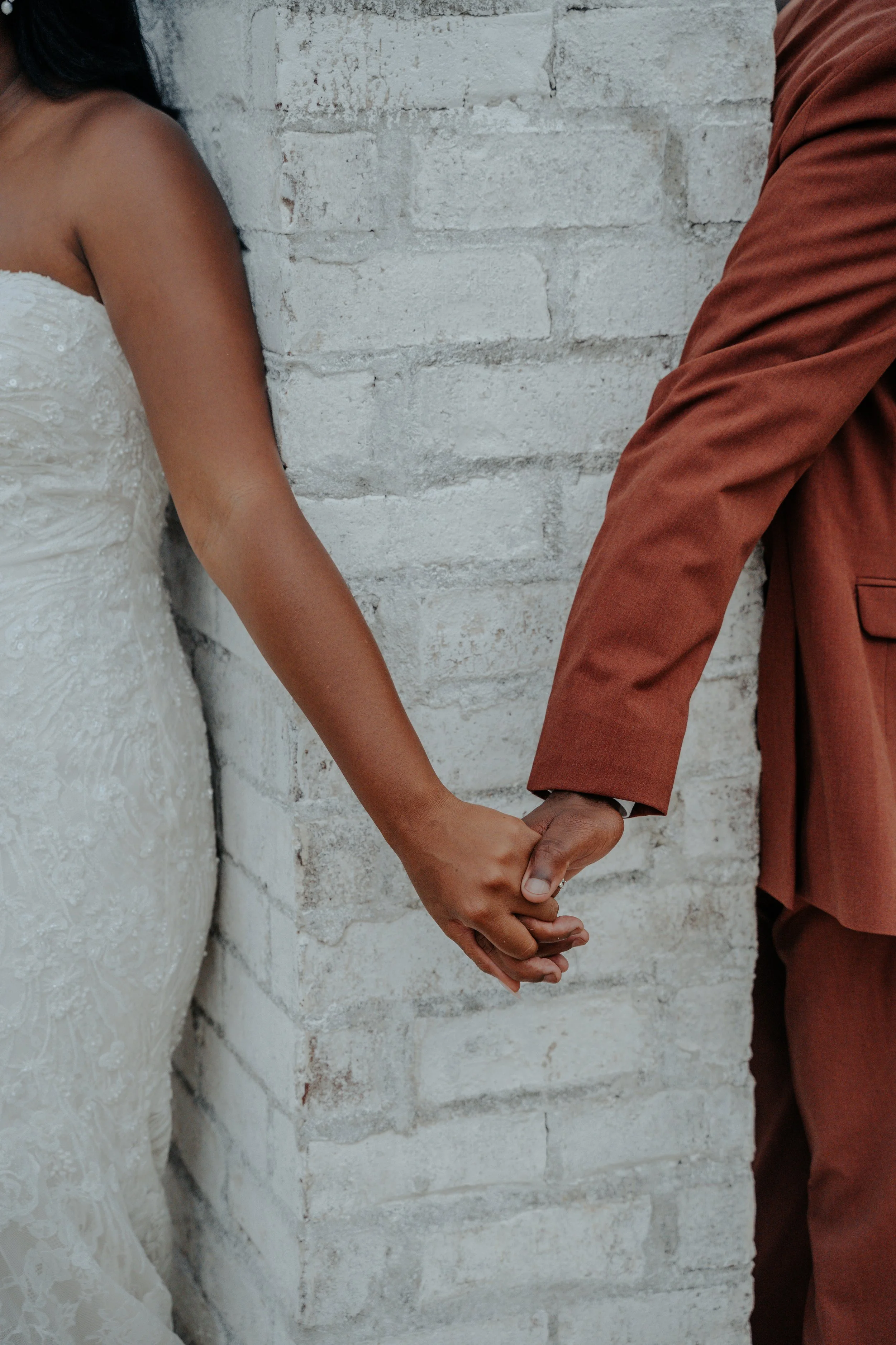 A couple holding hands, with the bride wearing a white lace wedding dress and the groom in a rust-colored suit, standing against a light-colored brick wall.