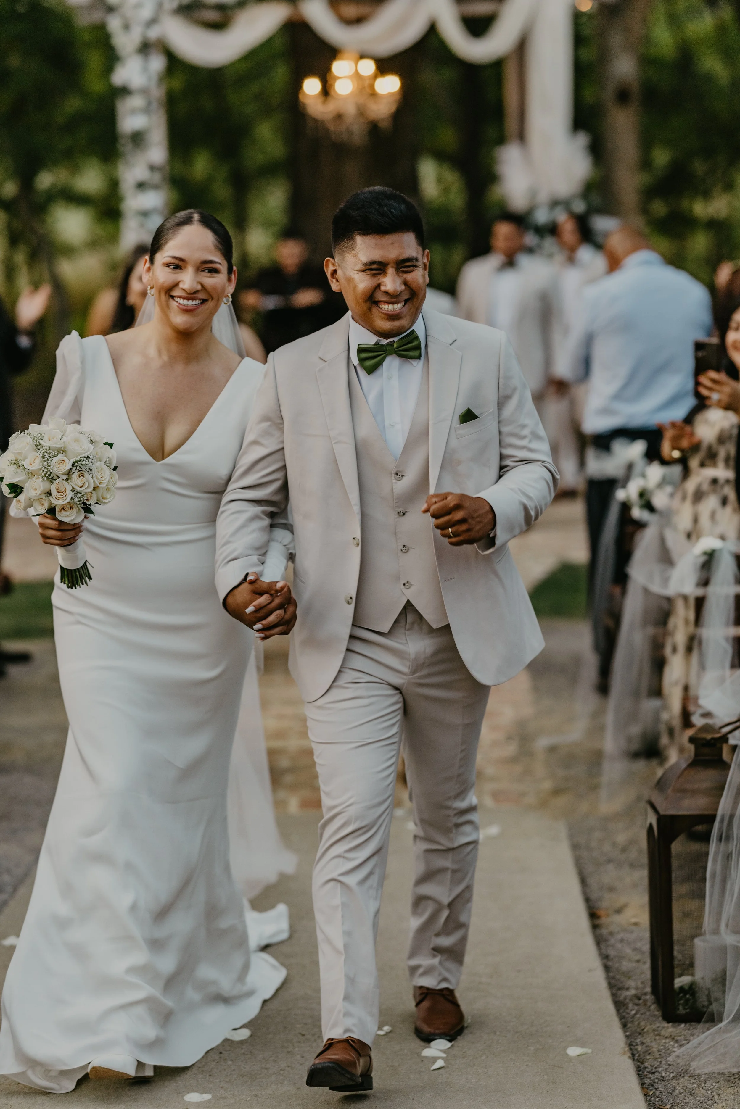 A newlywed couple walking down the aisle outdoors, smiling. The bride is wearing a white gown and holding a bouquet, and the groom is in a light suit with a green bow tie.