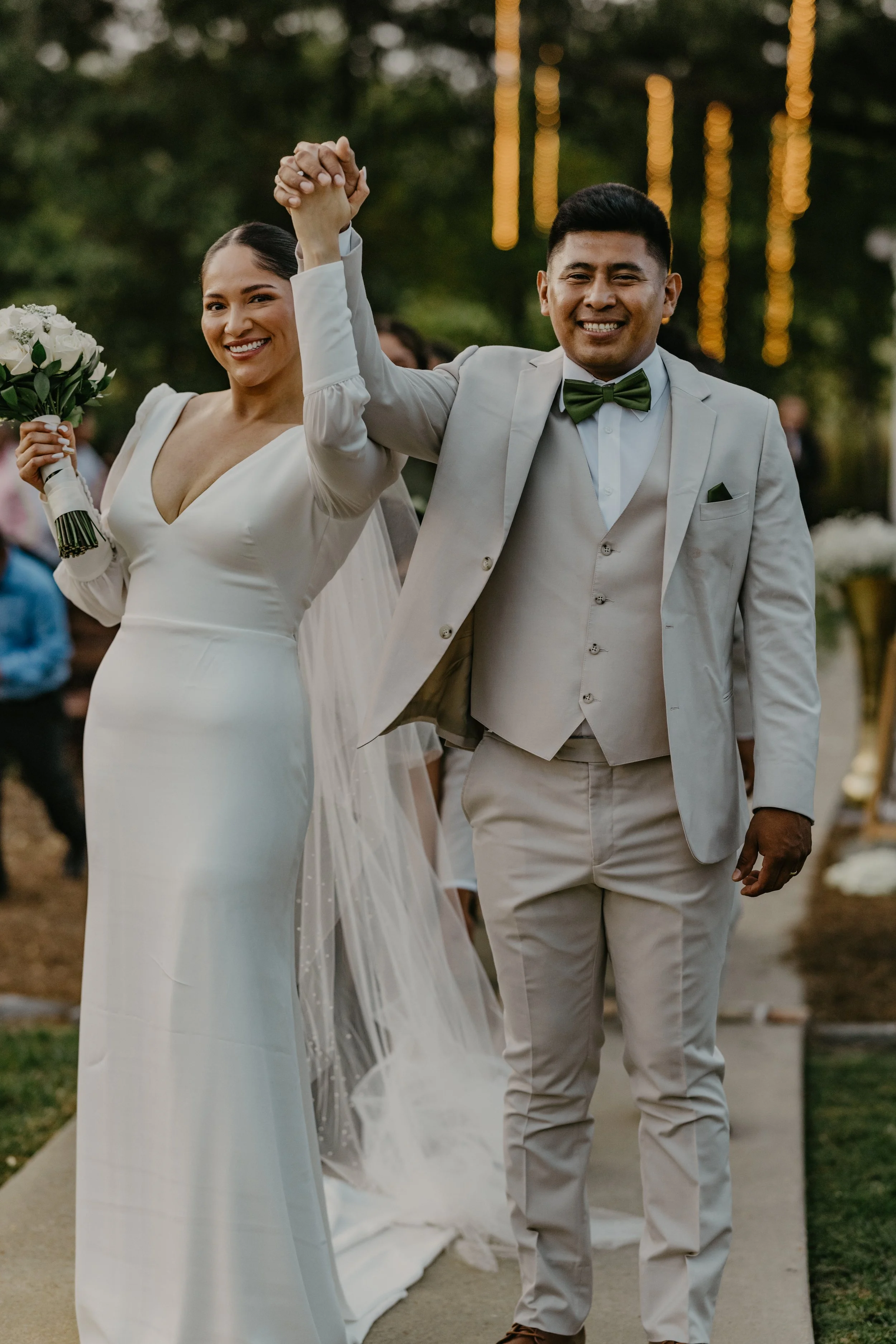 Bride and groom holding hands, smiling, walking outdoors at sunset, wedding attire, bride with bouquet, surrounded by guests and decorated trees.