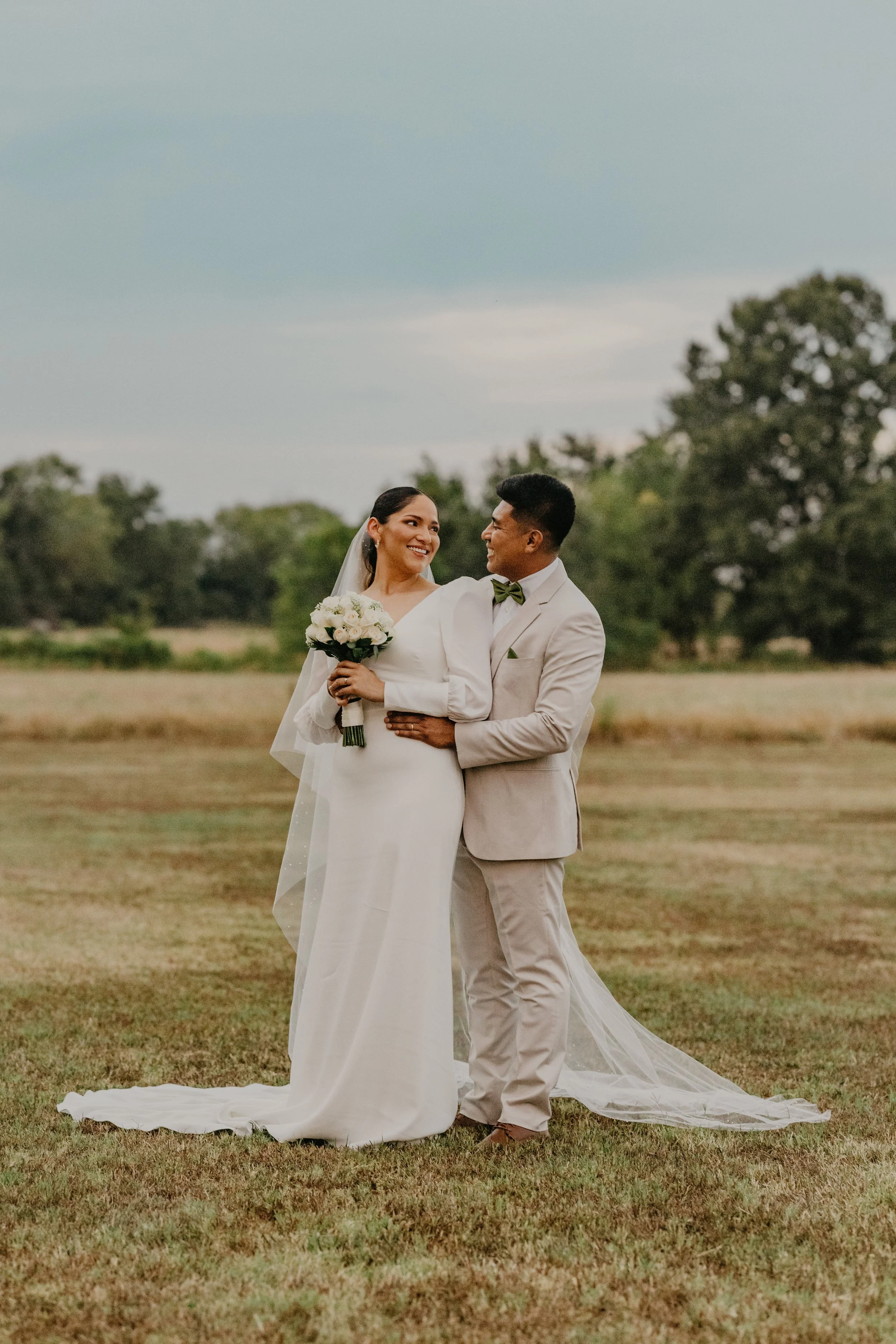 A bride and groom standing outdoors in a grassy field, smiling and looking at each other. The bride is holding a bouquet of white roses and wearing a long white wedding dress with a veil. The groom is dressed in a light-colored suit with a bow tie. T