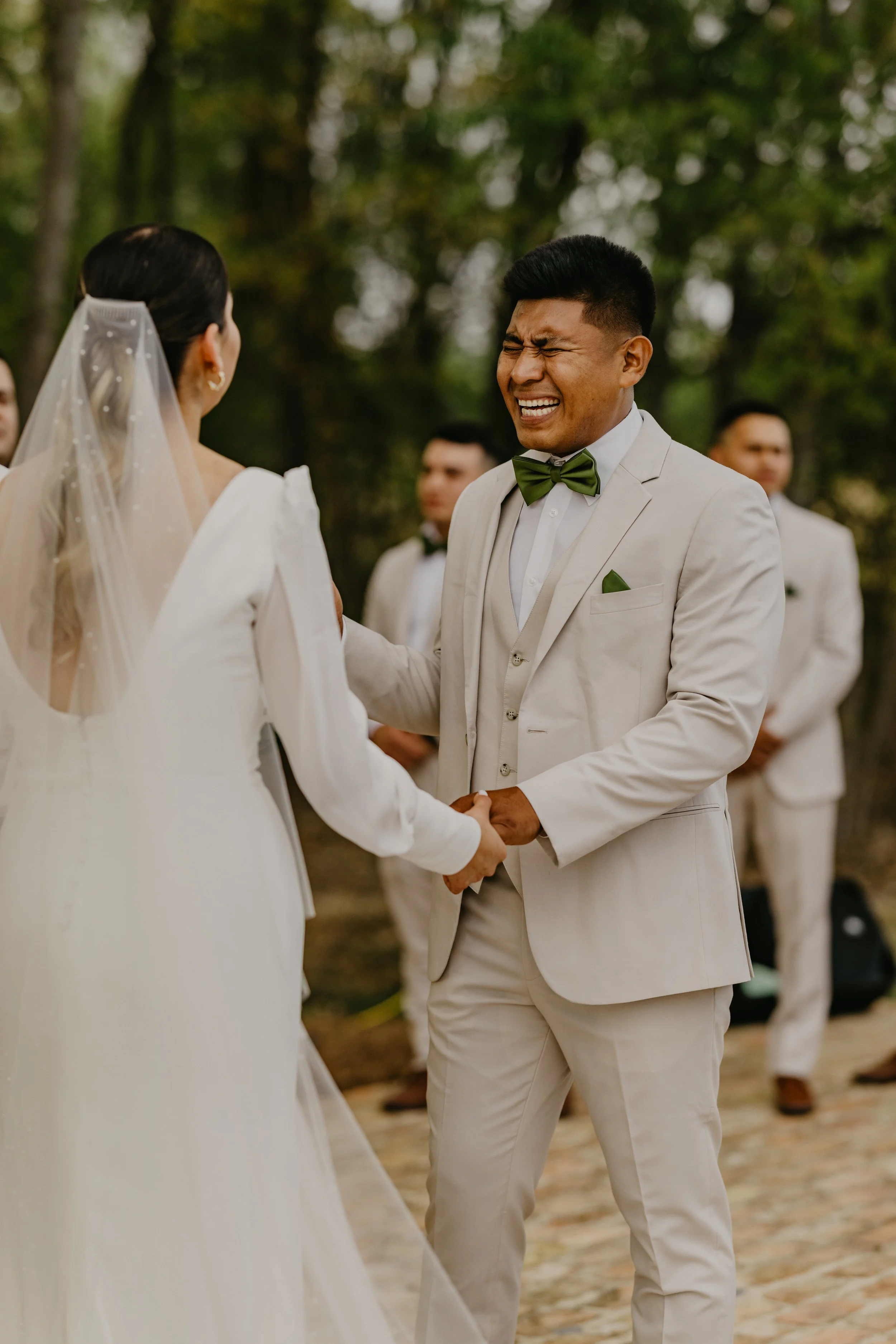 A groom in a beige suit and green bow tie is smiling with eyes closed while holding hands with a bride dressed in white at an outdoor wedding ceremony.