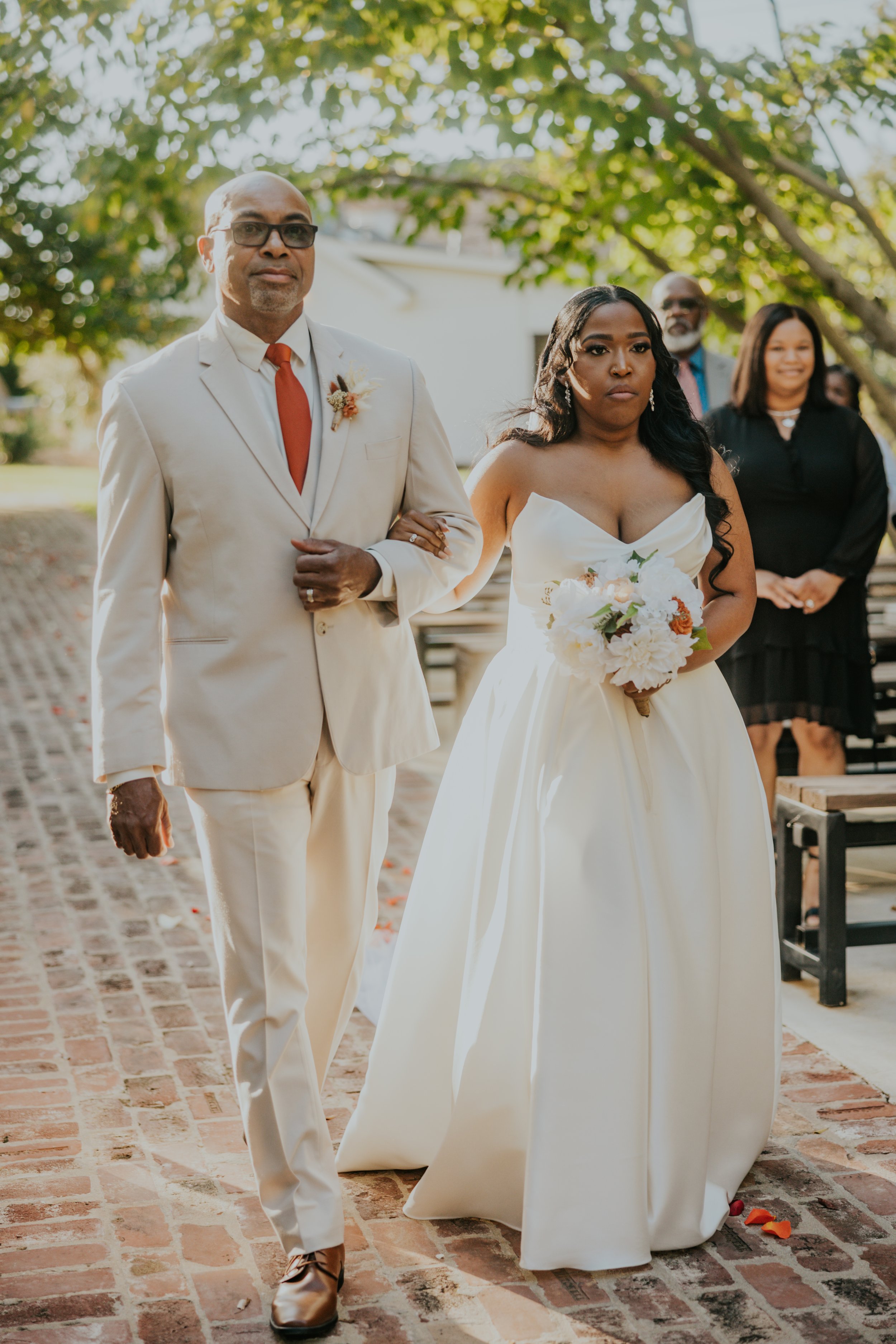 A bride walking down the aisle with a man, possibly her father, holding her arm. The bride is wearing an off-shoulder white wedding dress and holding a bouquet of flowers. The man is dressed in a light-colored suit with a red tie. In the background, 