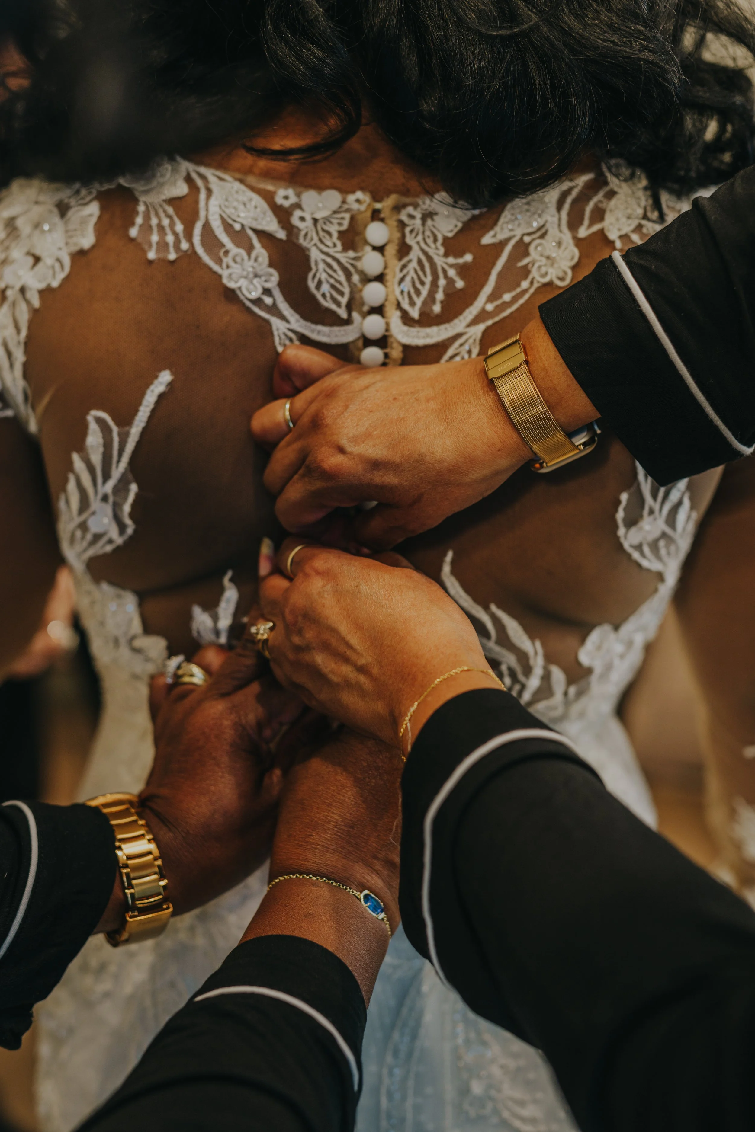Close-up of a bride in a lace wedding dress being assisted with the buttons by multiple people wearing watches and rings.