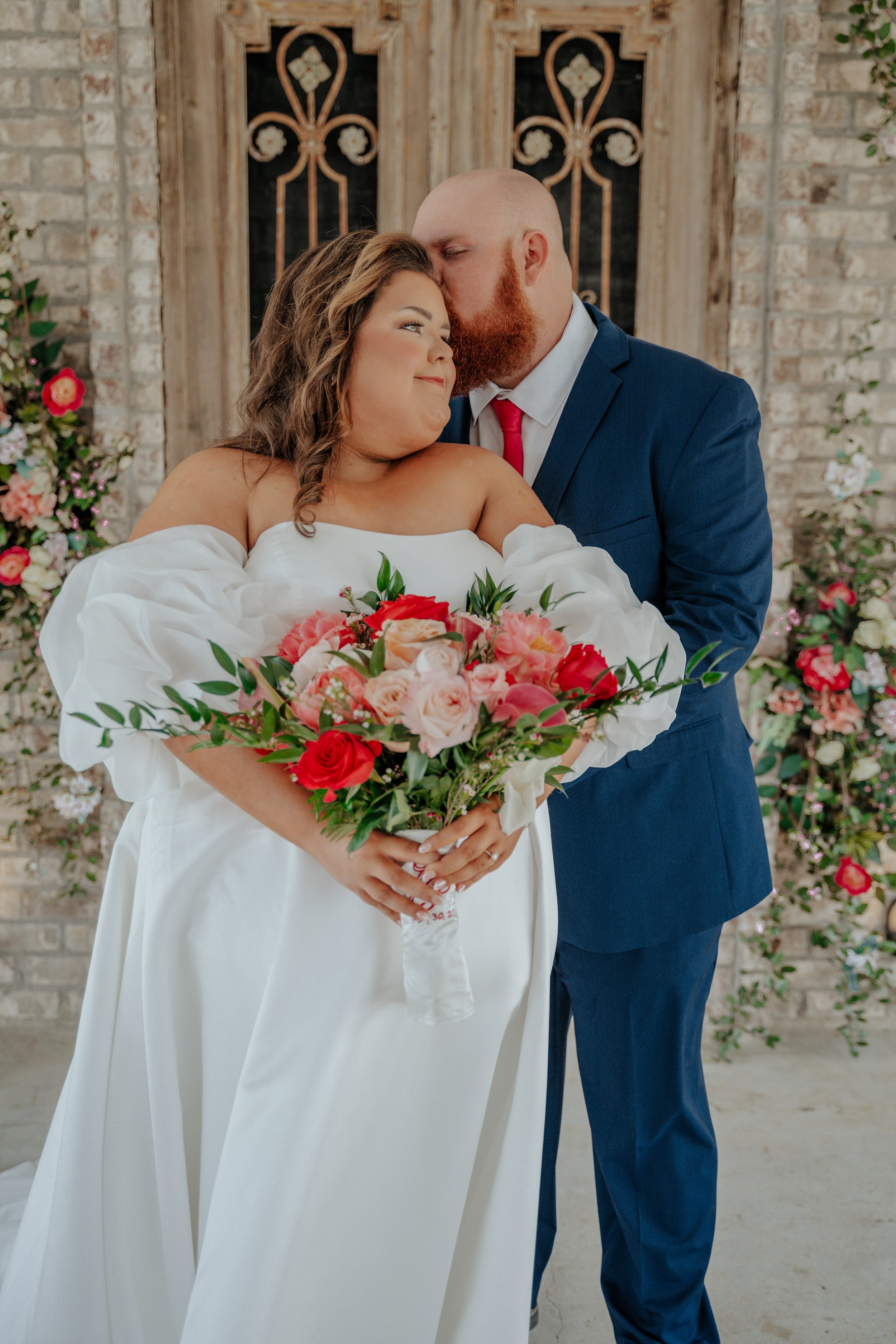 A bride and groom sharing a moment at their wedding, with the groom kissing the bride's forehead while she holds a bouquet of pink and red flowers.