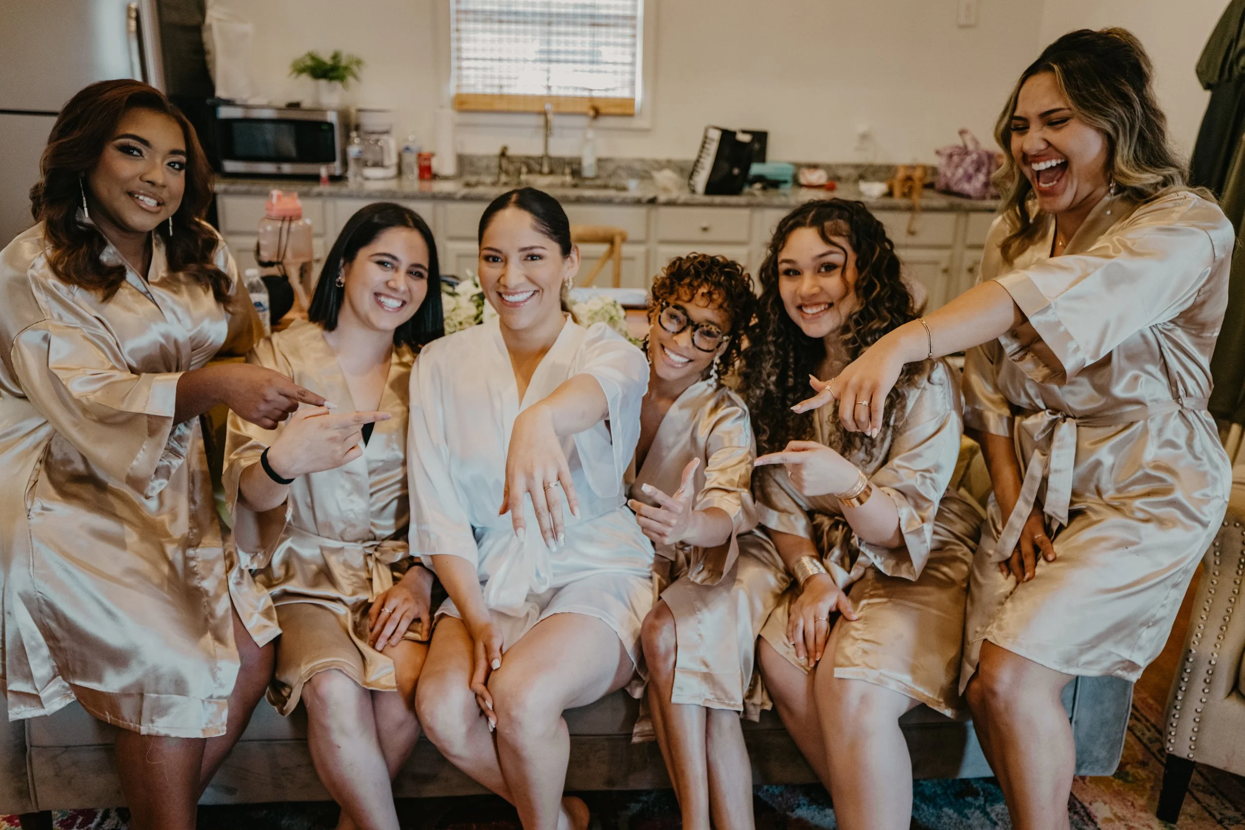 Group of women wearing matching satin robes, sitting and pointing at their rings, celebrating a wedding or special event in a cozy kitchen.