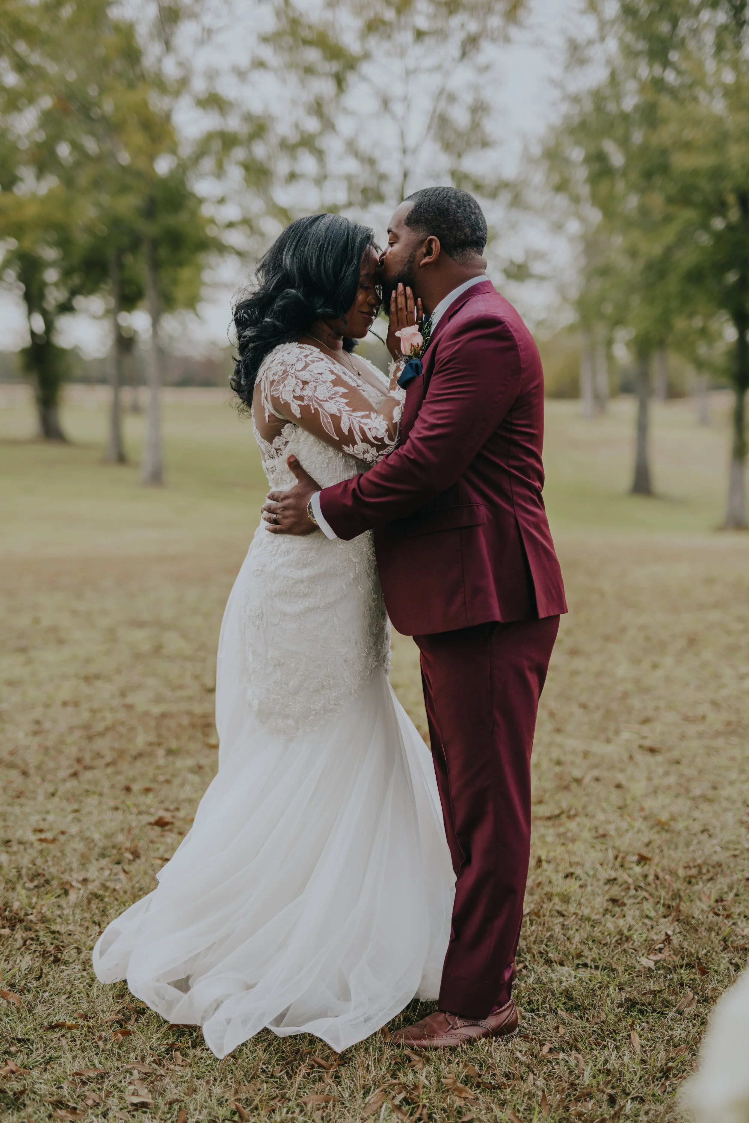 A bride and groom share a kiss in a park, with the groom kissing the bride's forehead and the bride holding his face gently.