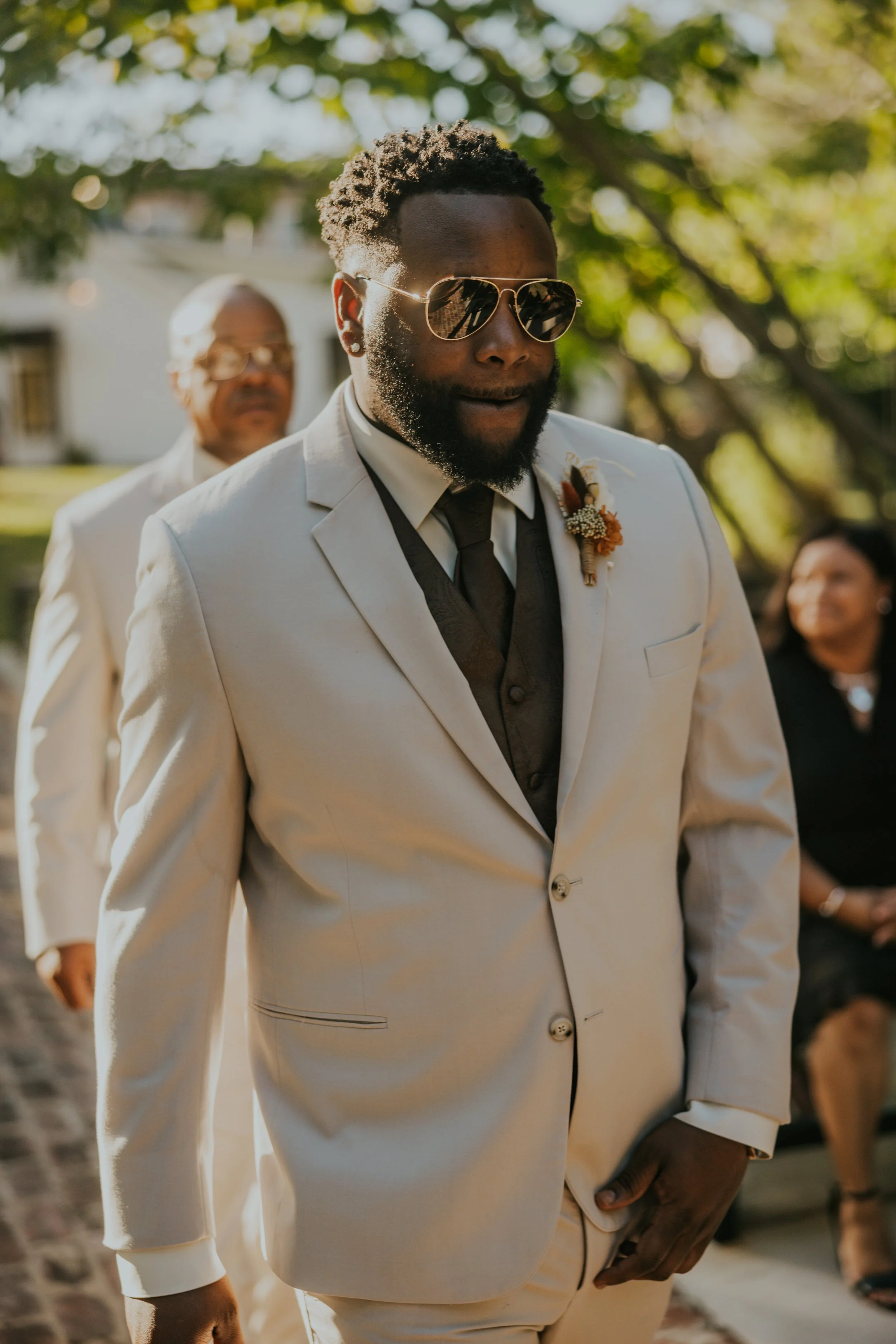A man in a cream suit, black tie, and sunglasses is walking outdoors during a wedding ceremony, with a boutonniere on his lapel and a woman seated in background.