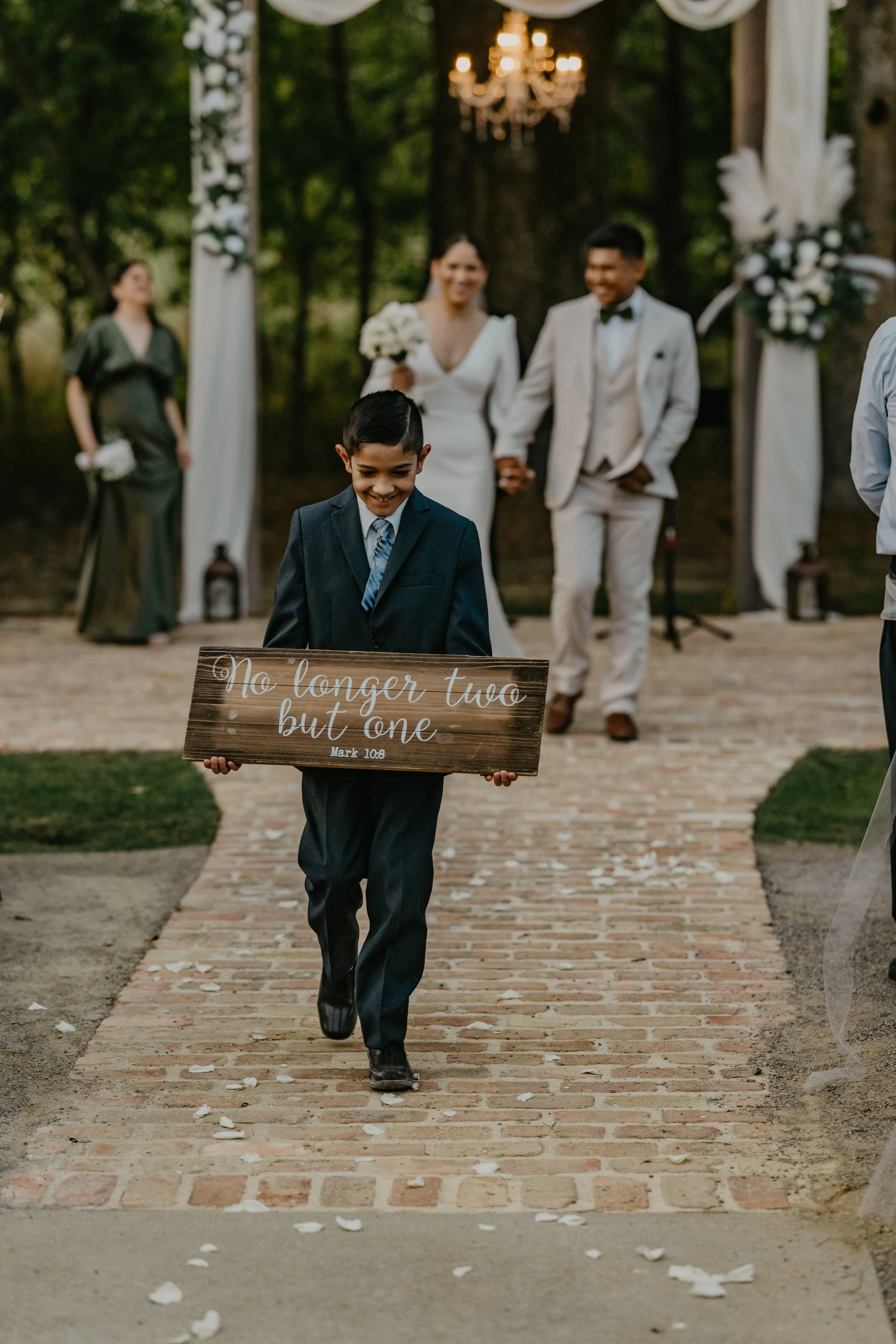A young boy in a suit walking down a brick aisle holding a wooden sign that reads, 'No longer two but one, Mark 10:8'. In the background, a bride and groom are holding hands, smiling, with the bride holding a bouquet of white flowers. Flower arrangem