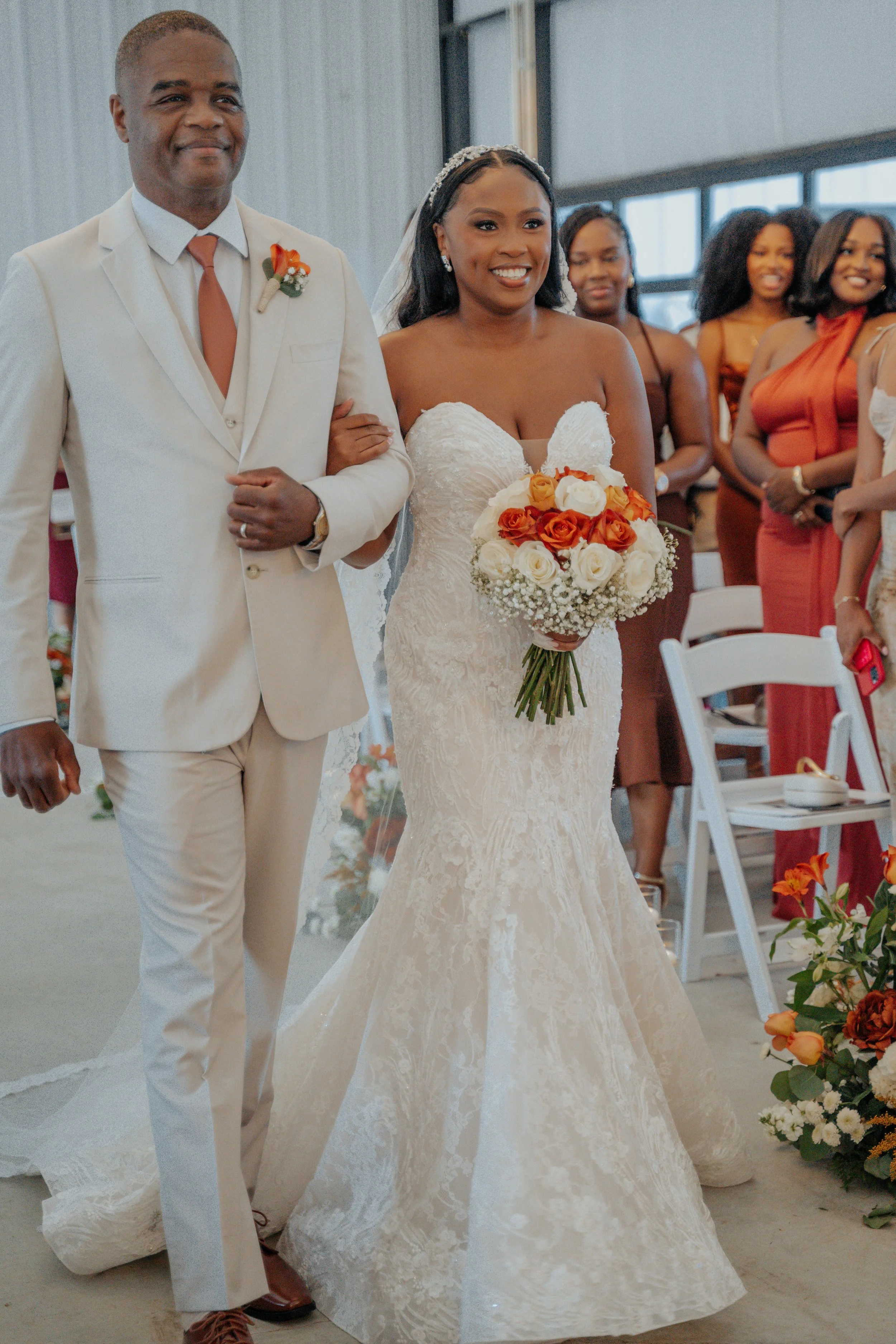 A bride walking down the aisle with her father at a wedding ceremony, holding a bouquet of orange and white roses, with bridesmaids in the background.