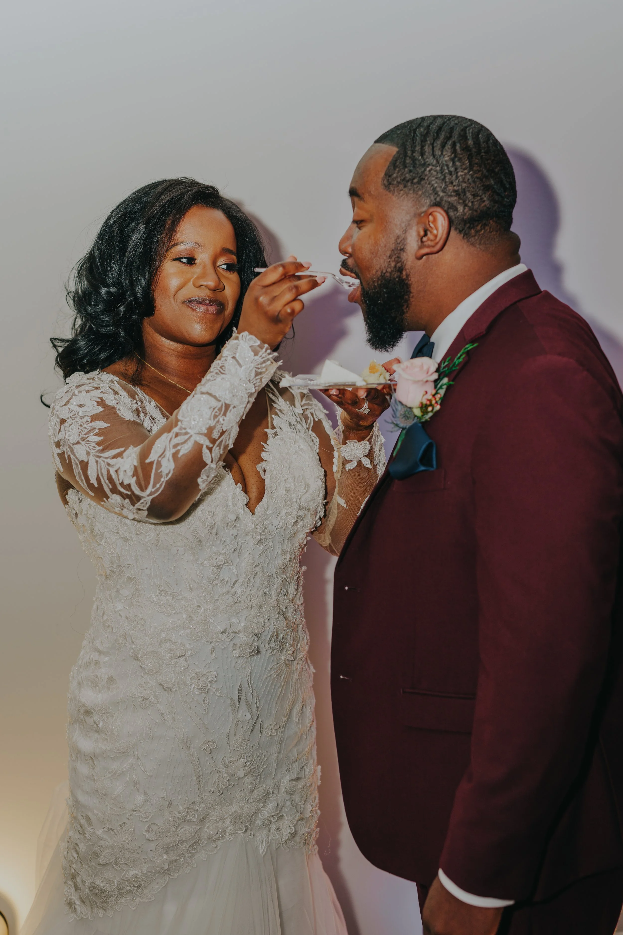 A bride in a white lace wedding dress feeding cake to a groom in a burgundy suit.