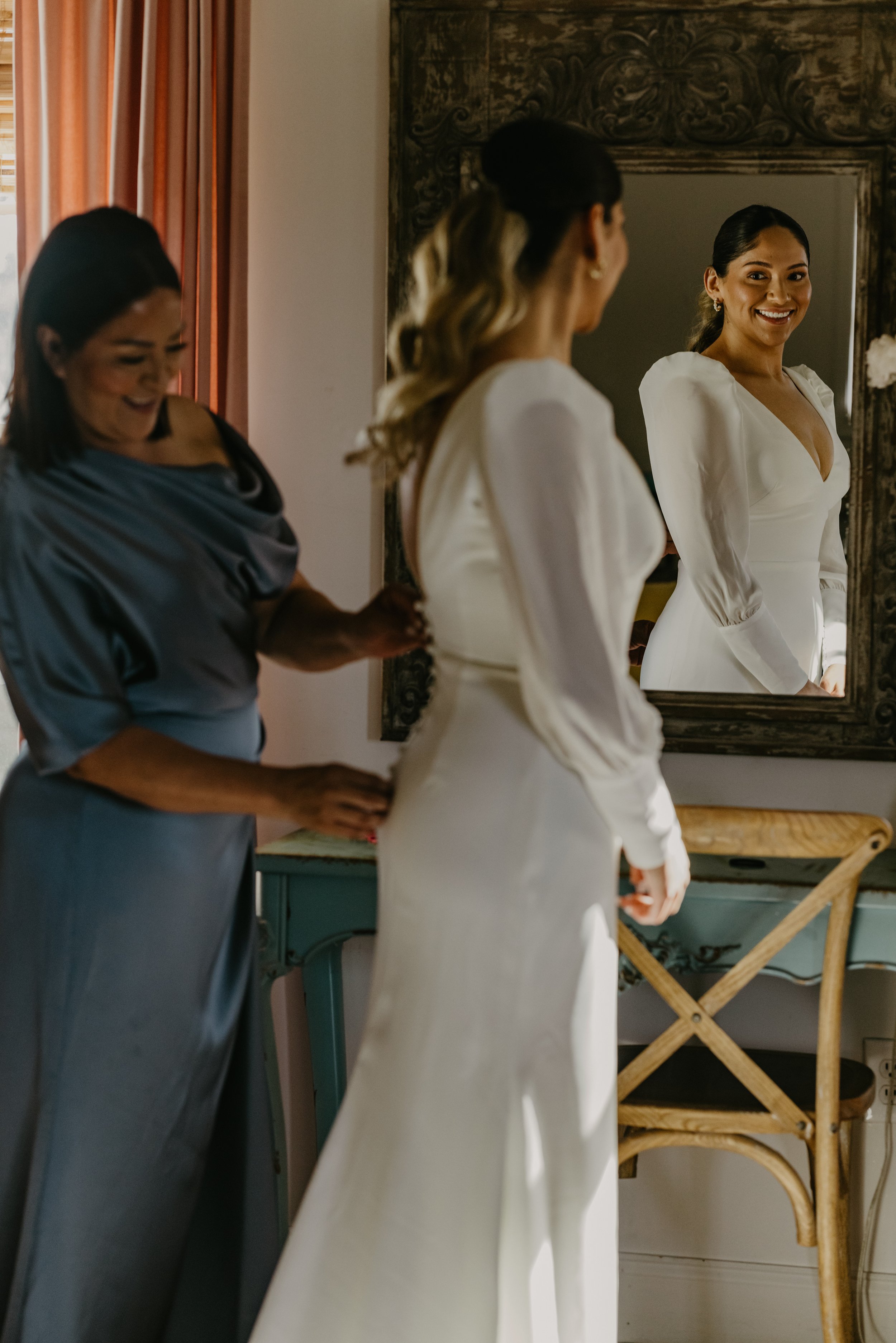 A bride in a white dress looks at her reflection in a mirror, smiling, while a woman helps her adjust her dress.