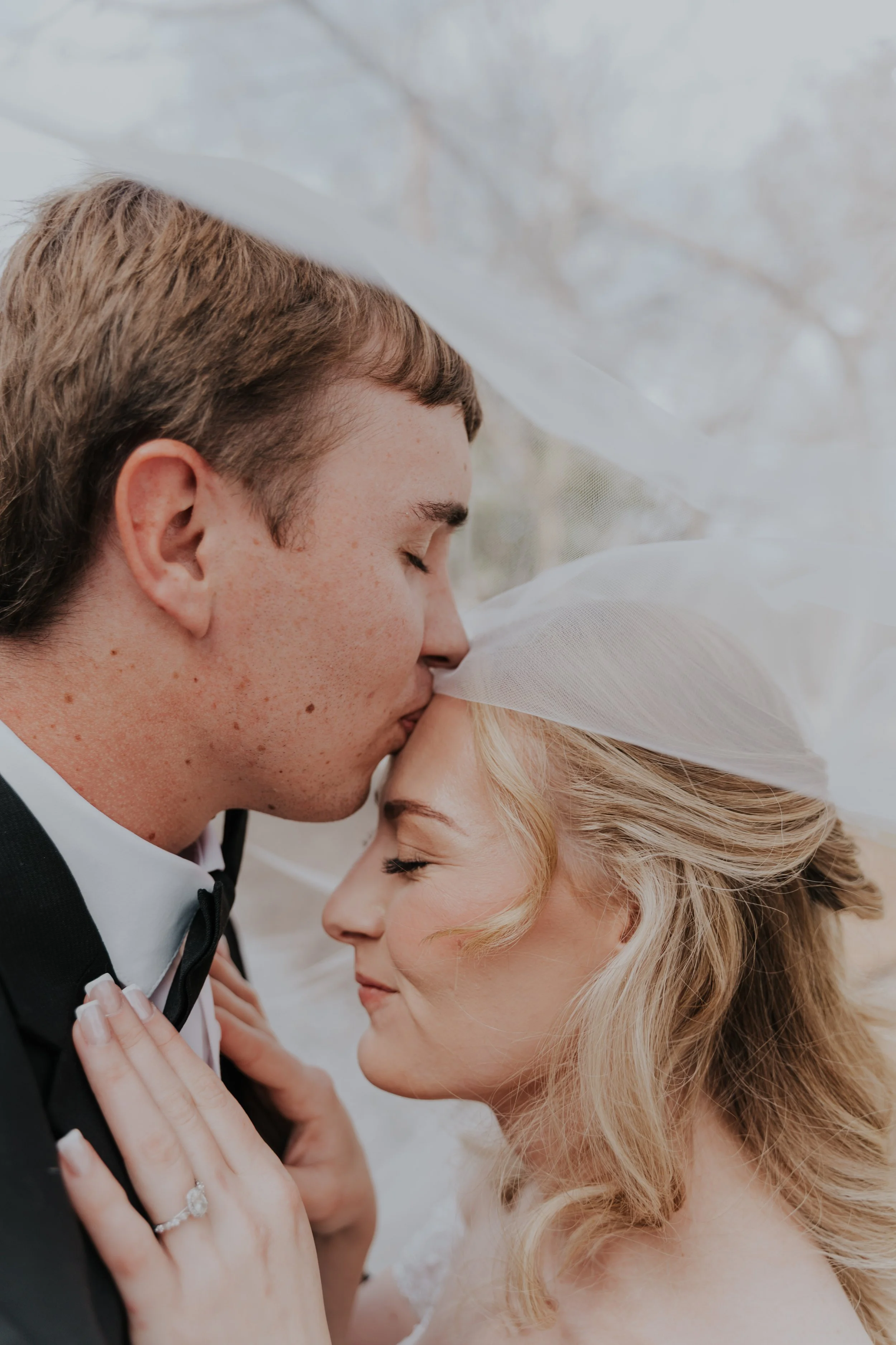 A bride and groom in wedding attire share an intimate moment with their foreheads touching, eyes closed, under a sheer veil.