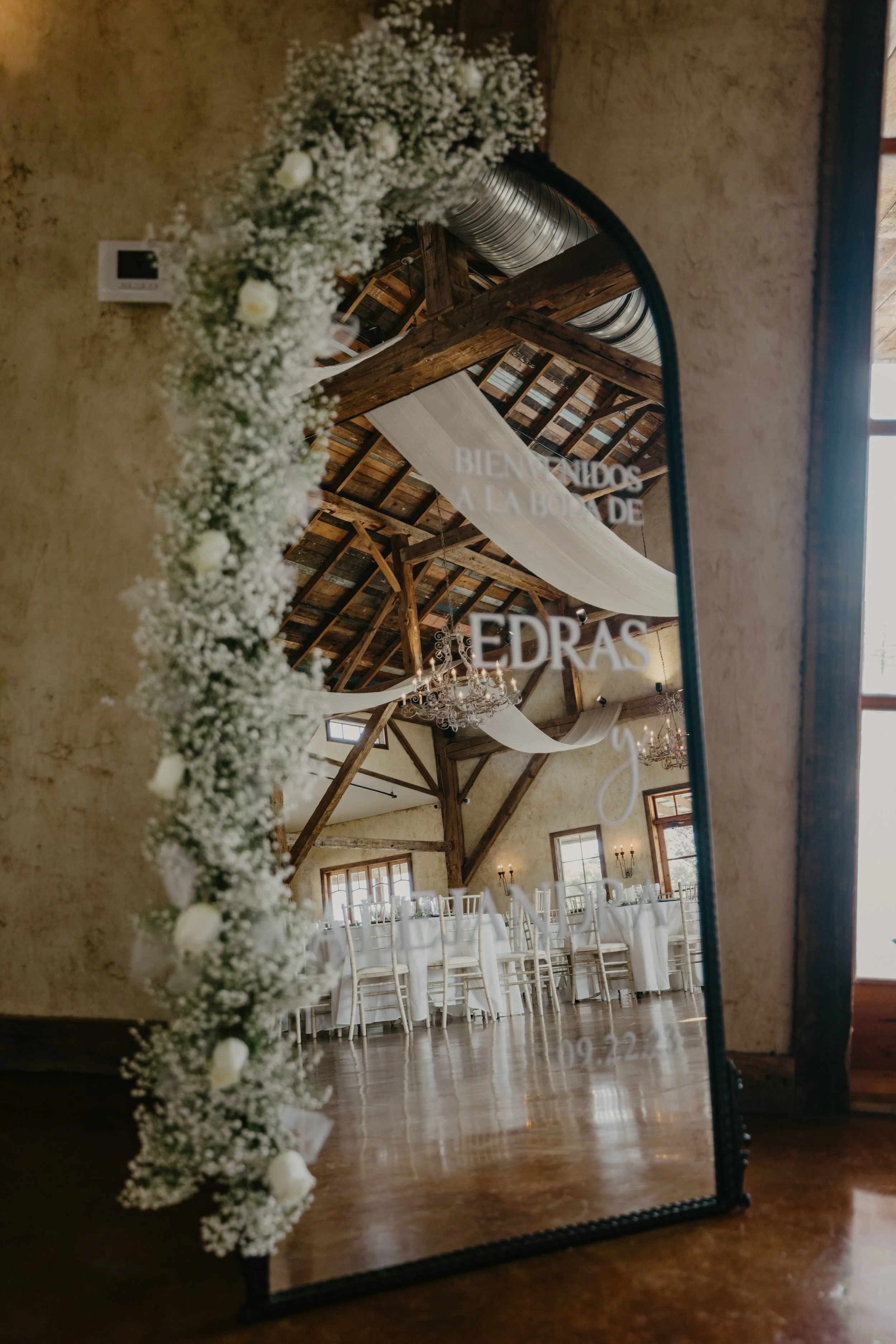 Wedding reception hall reflected in a large mirror decorated with white baby's breath flowers. The hall has wooden beams, chandeliers, and a sign that reads "Bienvenidos a la boda de Edra's."