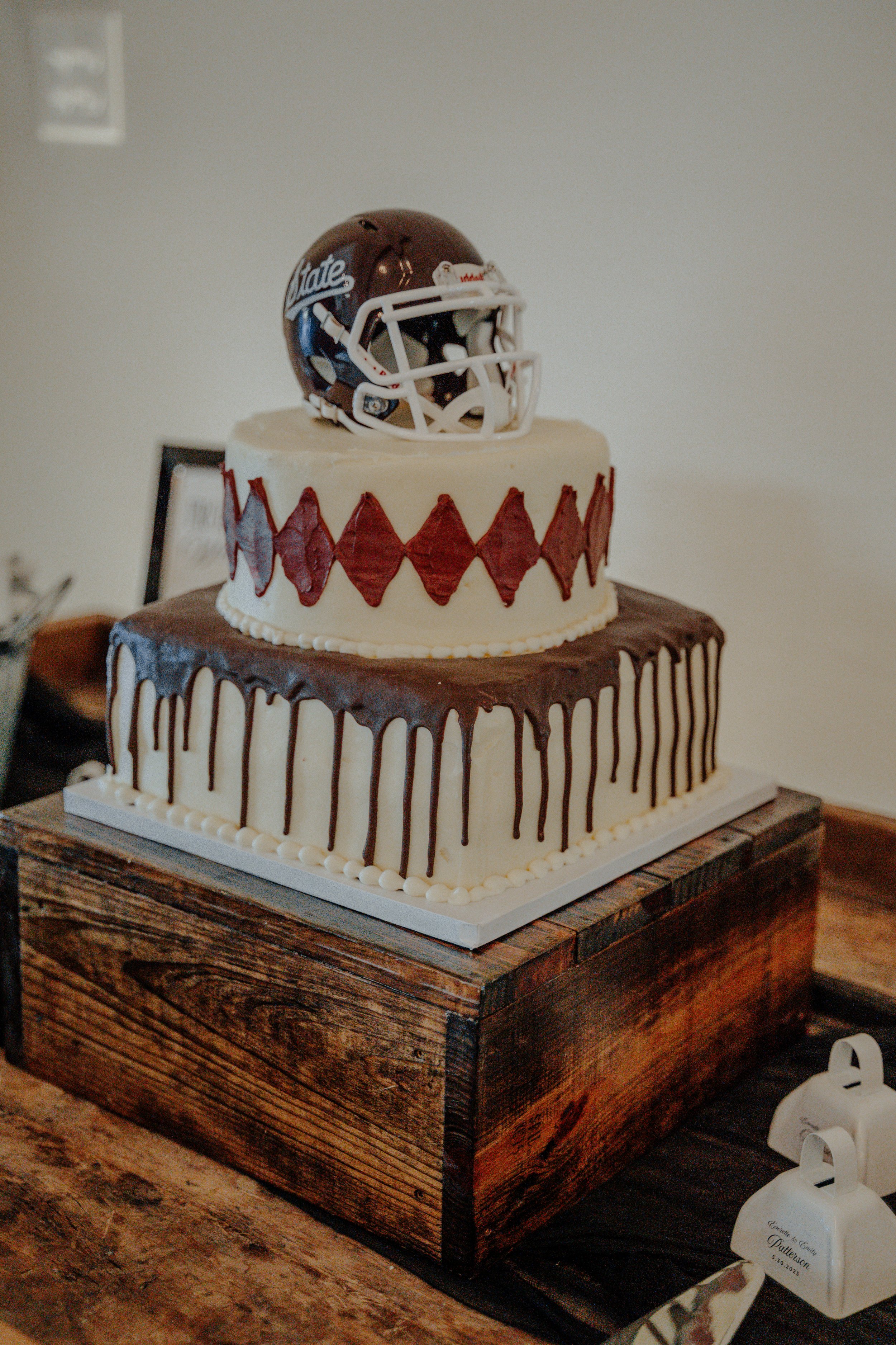 Two-tiered cake with football helmet decoration on top; the upper cake has a red diamond pattern, and the bottom cake has a chocolate drip glaze.