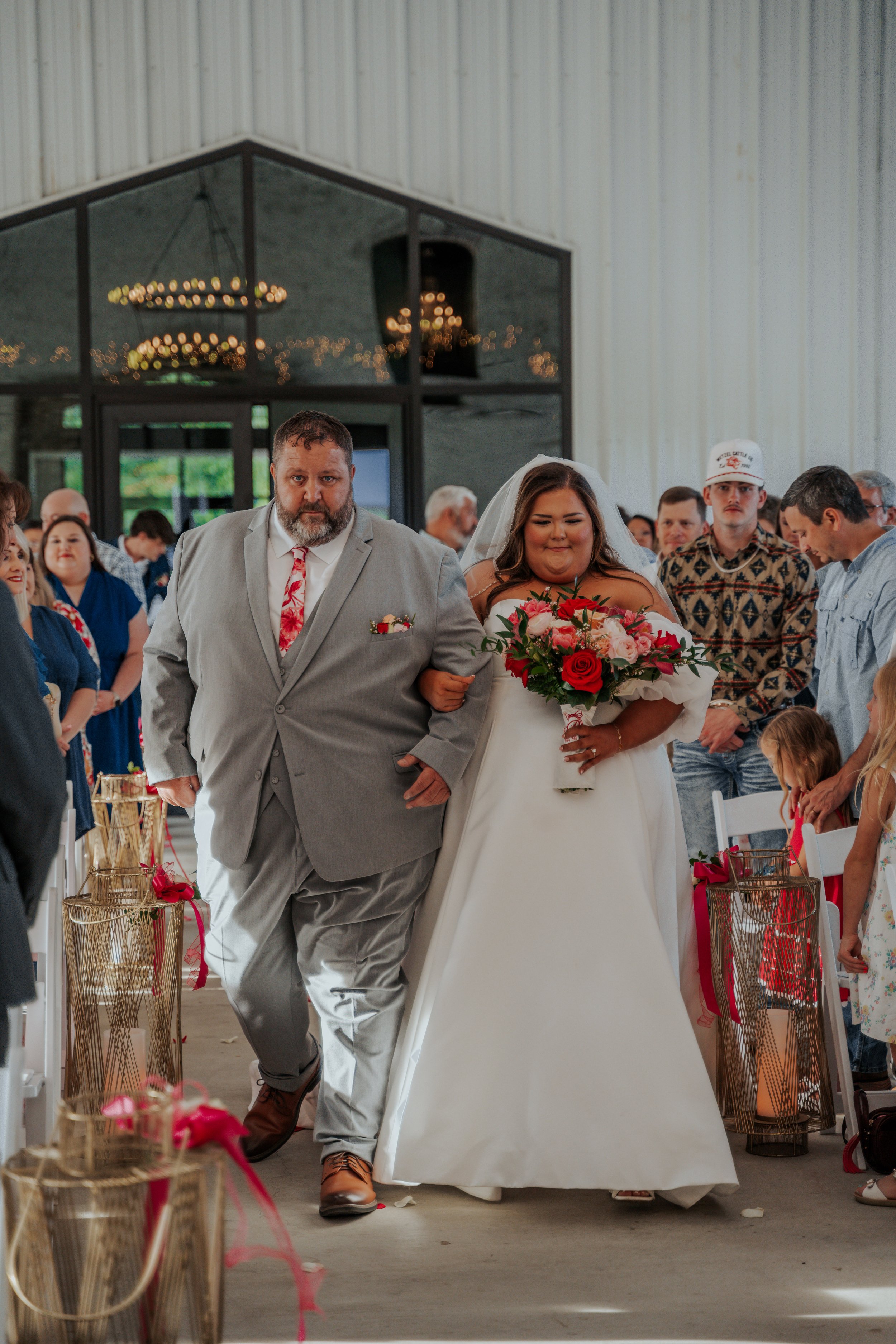 A bride walking down the aisle arm-in-arm with a man, likely her father, during a wedding ceremony inside a venue with guests seated on both sides. The bride is holding a bouquet of red and pink flowers and wearing a white wedding gown. The man is we