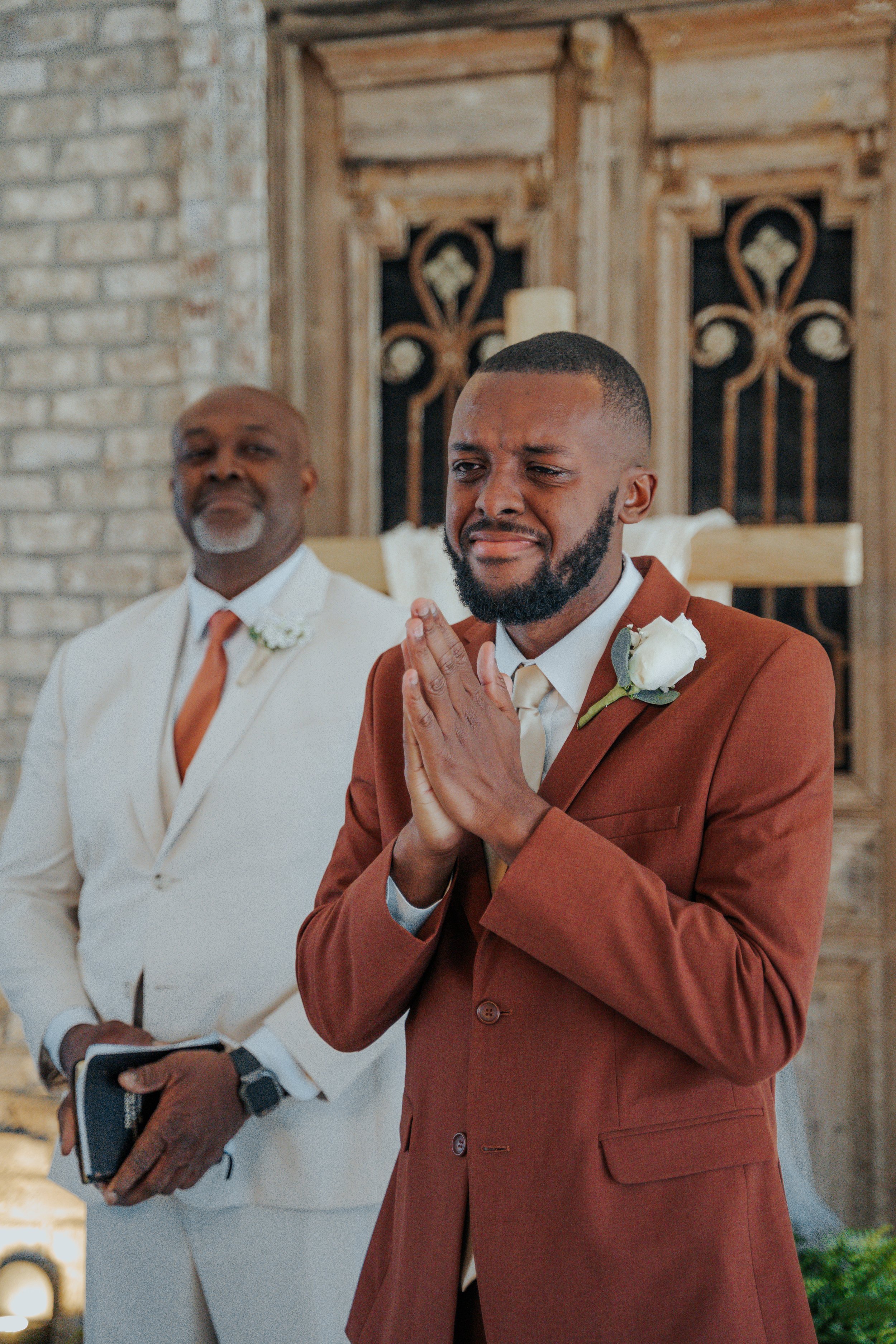 A groom in a brown suit with a white boutonniere prays during a wedding ceremony, with an officiant in a white suit standing behind him.
